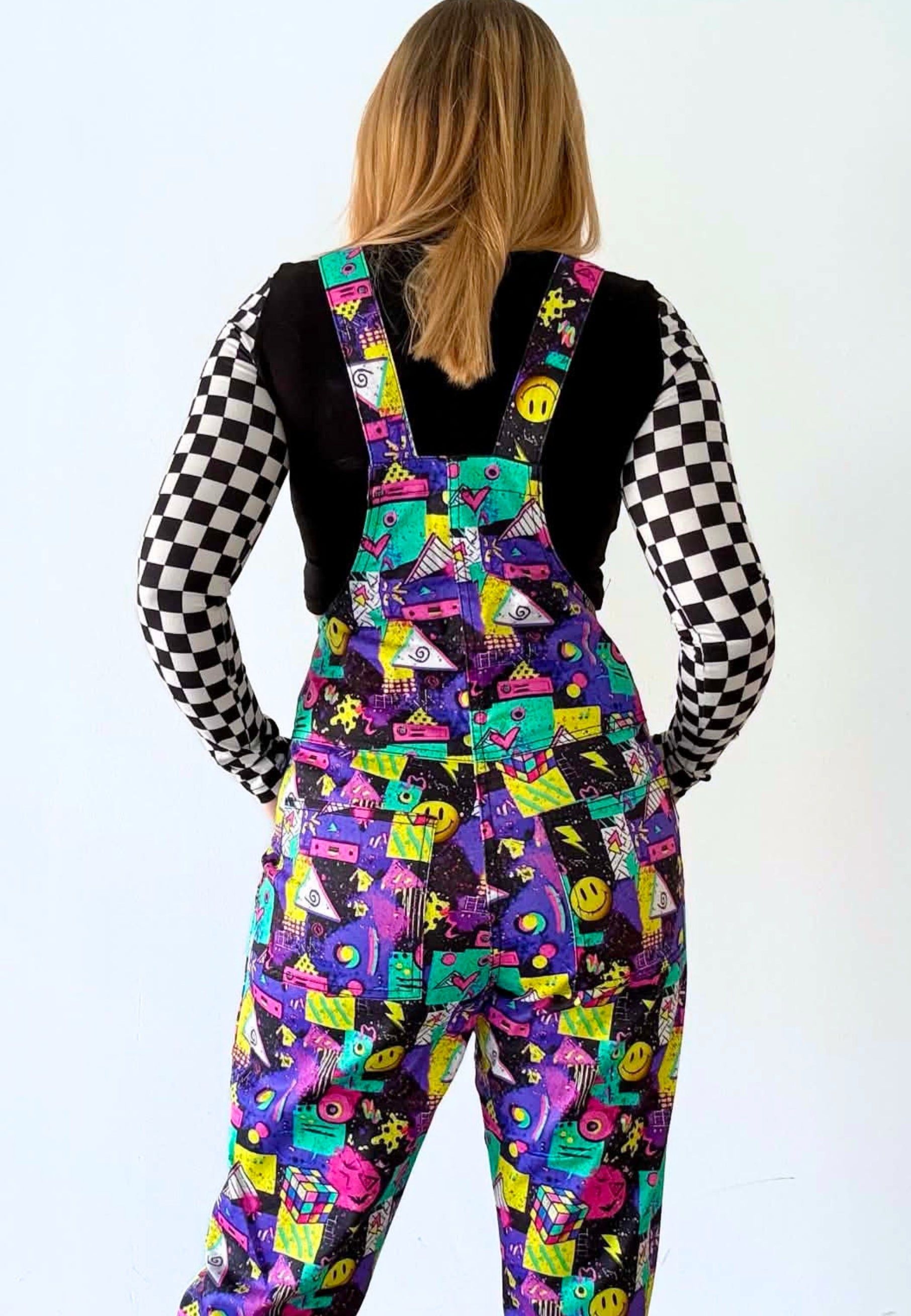 A person shown from the back wearing multicoloured 80s 90s patterned dungarees overalls over a black vest and a black and white checkered long-sleeve shirt, against a plain white background.