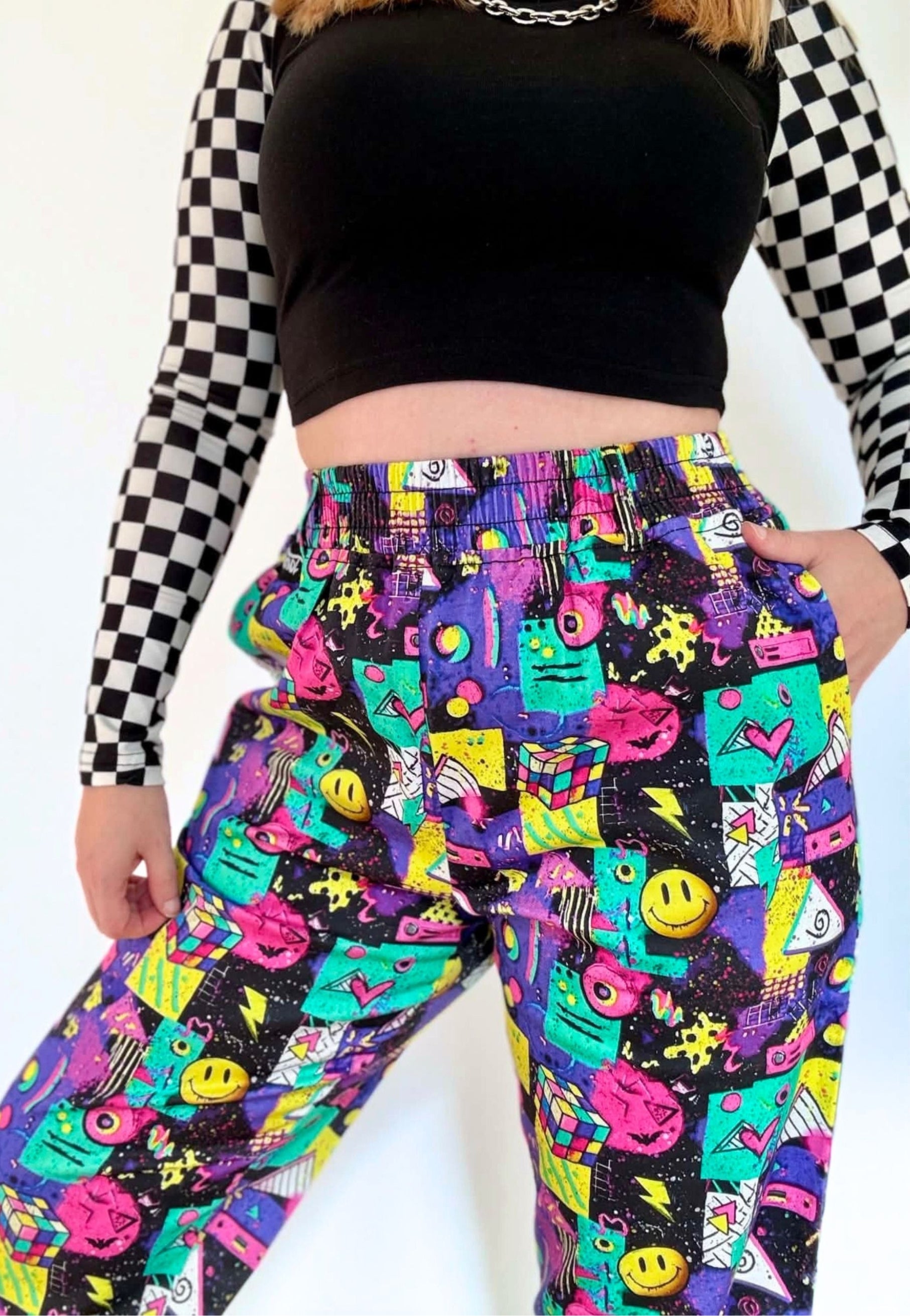 A person wearing high-waisted, wide-l leg pants with a vibrant multicoloured 80s 90s print of smiley faces, neon shapes, and retro blocks on a black background, styled with a black checkered long sleeve top and a black crop top.