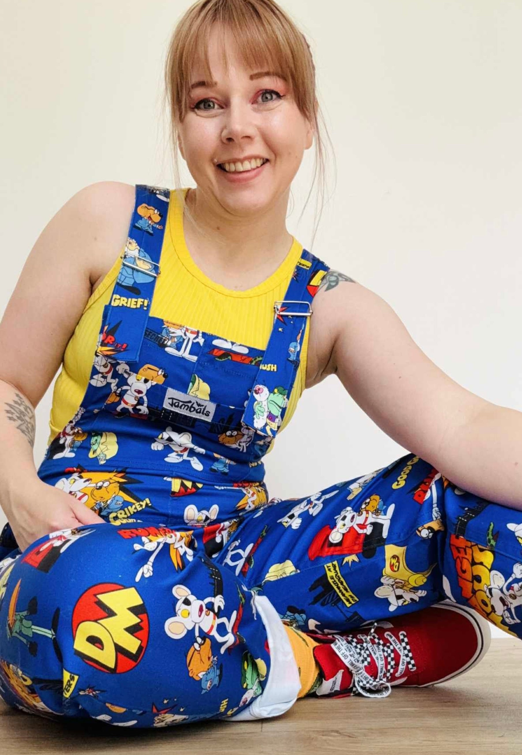 Woman sitting down wearing blue Danger Mouse dungarees with a yellow top underneath. Studio photo