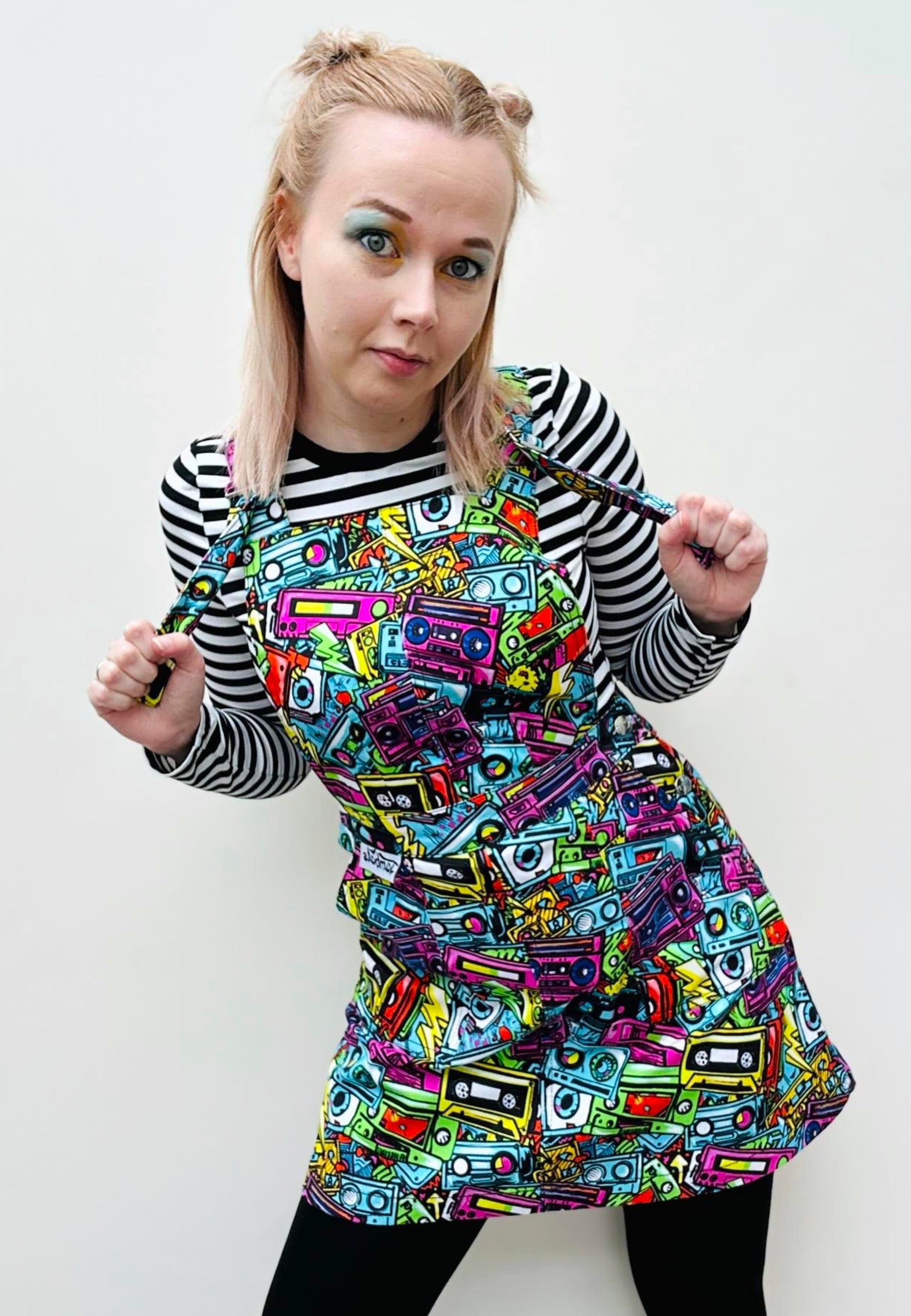 A woman wearing a vibrant multicolored pinafore dress with a vibrant street art style print, featuring retro 80's 90's cassette motifs. 