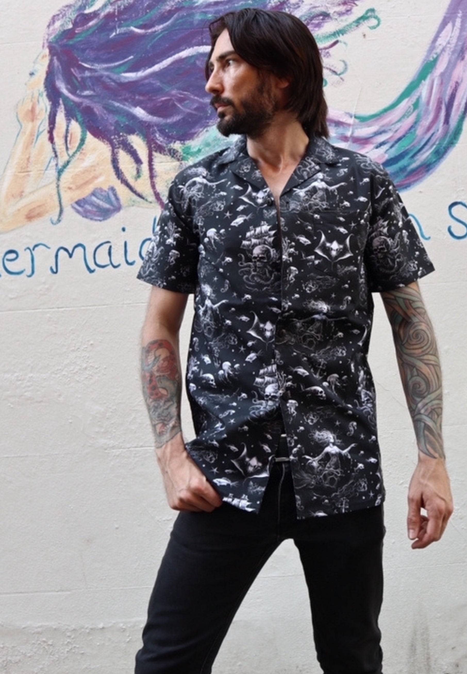 Man wearing a black shirt with a gothic under the sea print..ghostly mermaids and pirate ships. Man is standing in front of a mermaid wall mural.