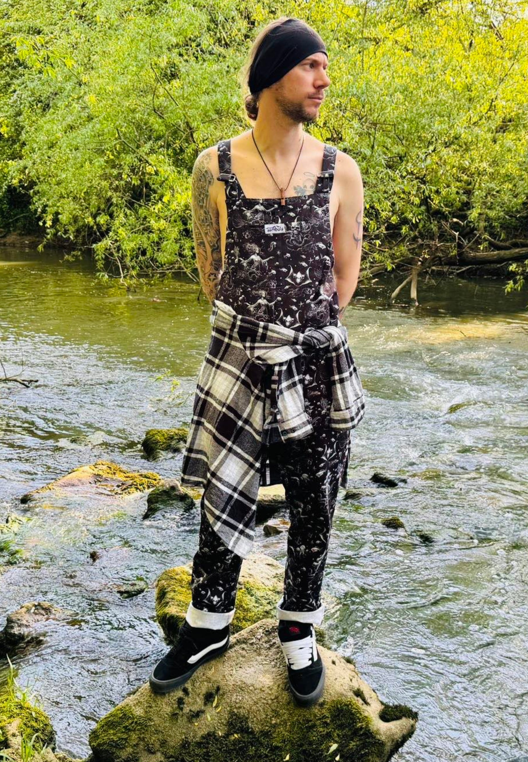 A man wearing black dungarees with a pattern of ghostly pirate skull ships and eerie sirens. Outdoors stood on a rock in the middle of a stream. 