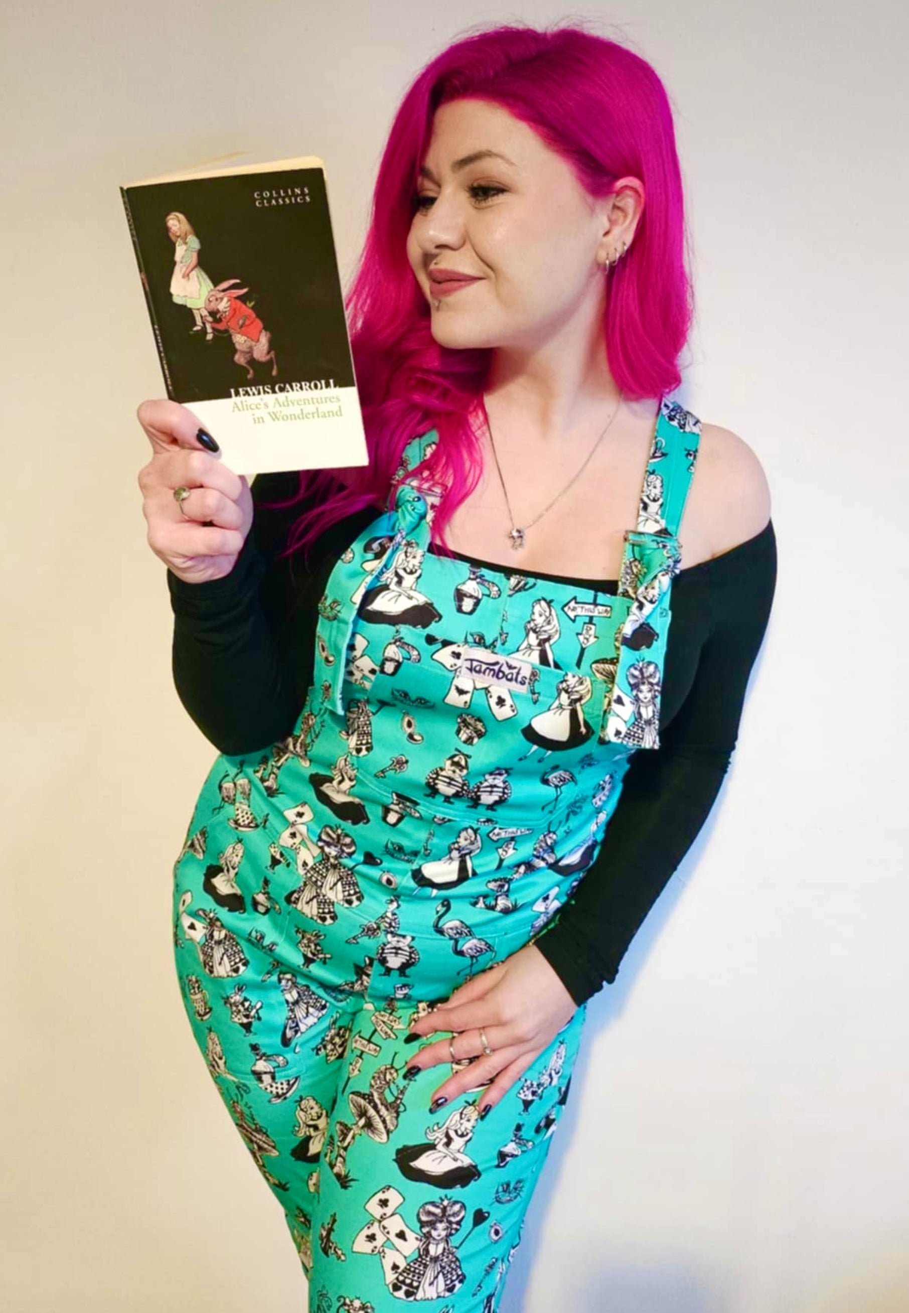 A woman with beautiful deep pink hair I’d wearing Alice in Wonderland print dungarees with black and white characters, standing outdoors.  Reading an Alice in Wonderland Book 