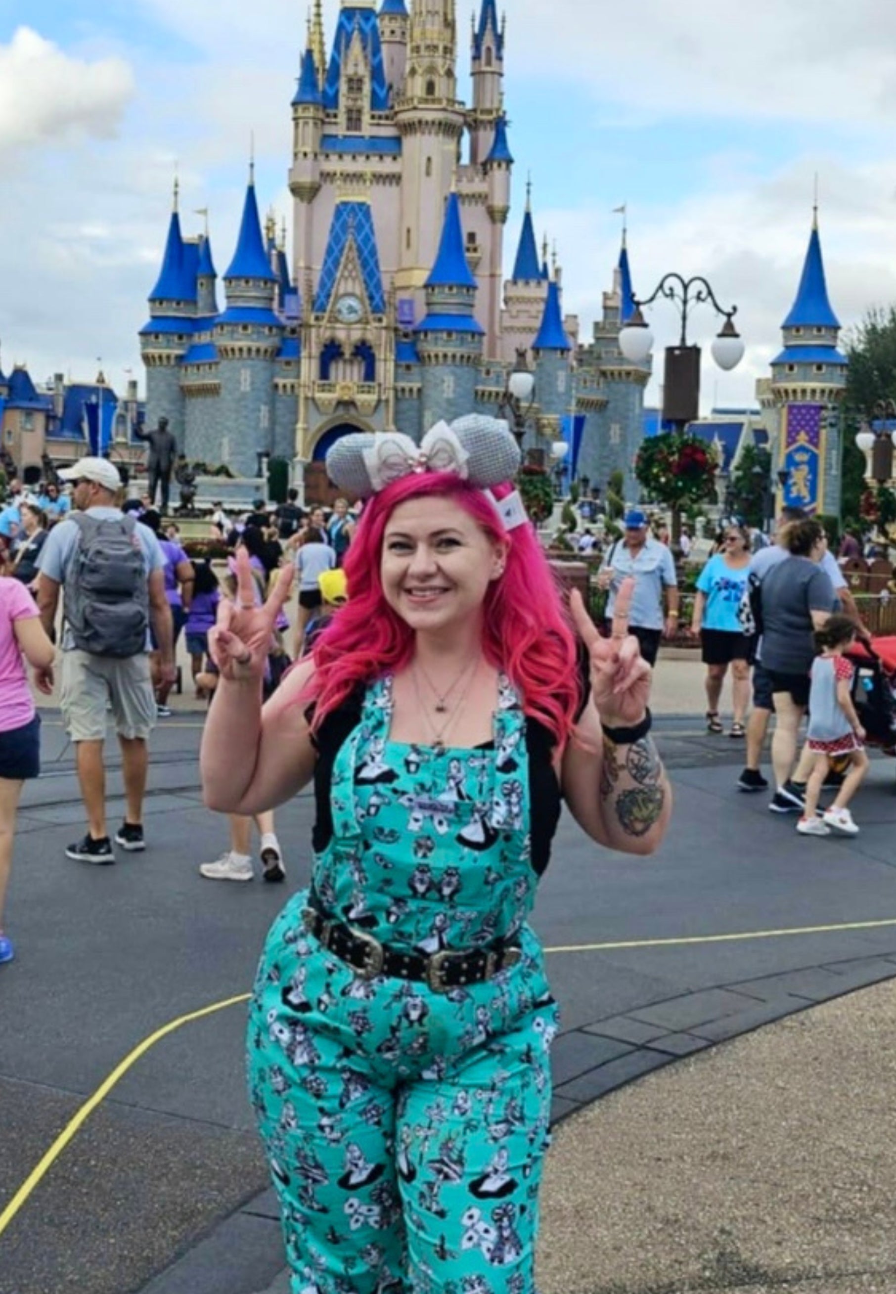 A woman with beautiful deep pink hair wearing Alice in Wonderland print dungarees with black and white characters, standing outdoors.