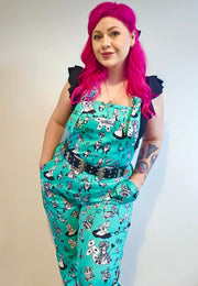 A woman with beautiful deep pink hair I’d wearing Alice in Wonderland print dungarees with black and white characters, standing outdoors. 