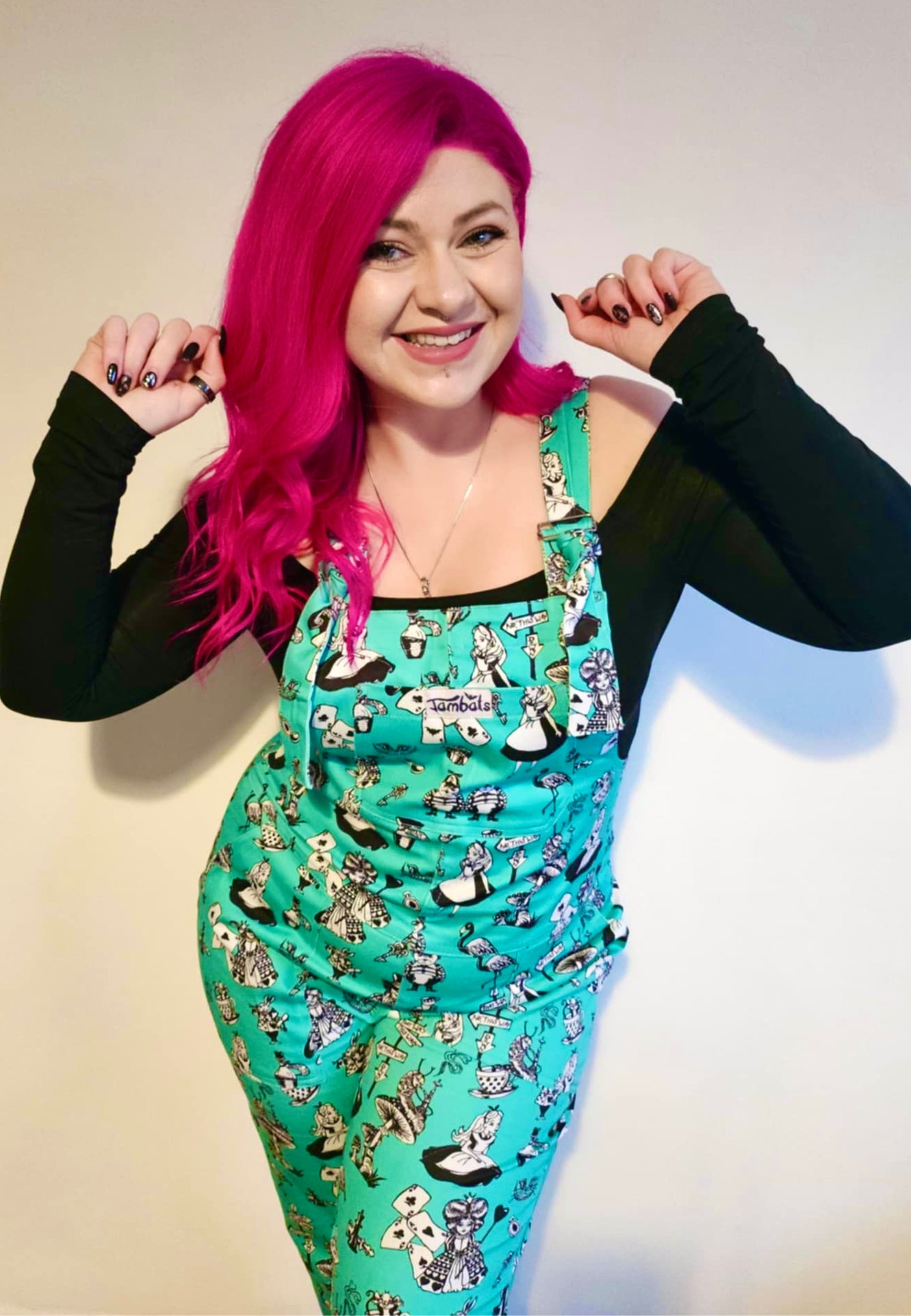 A woman with beautiful deep pink hair wearing Alice in Wonderland print dungarees with black and white characters. 