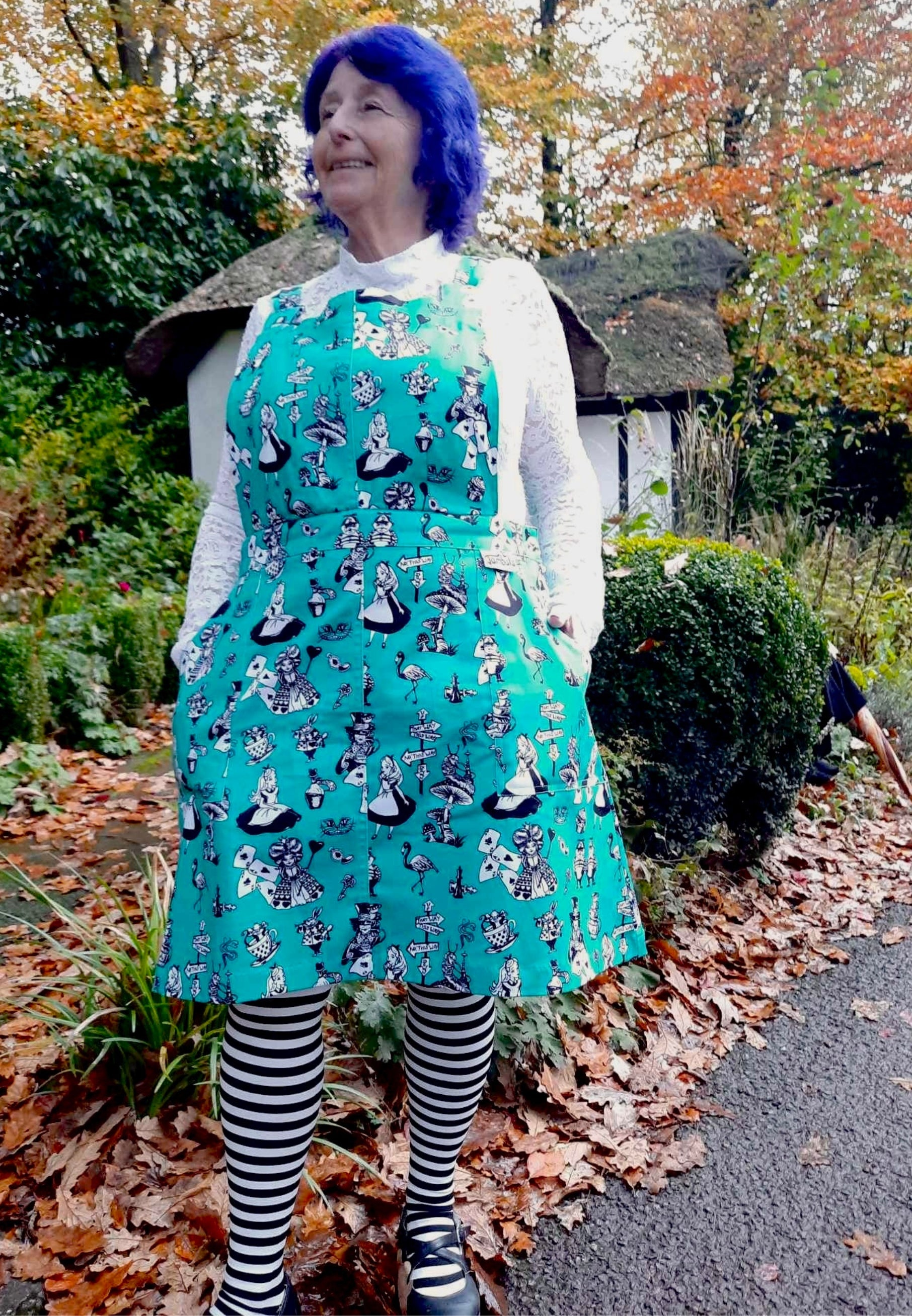 A woman wearing an Alice in Wonderland print pinafore dress with black and white characters, standing outdoors. Also wearing quirky black and white striped tights. 