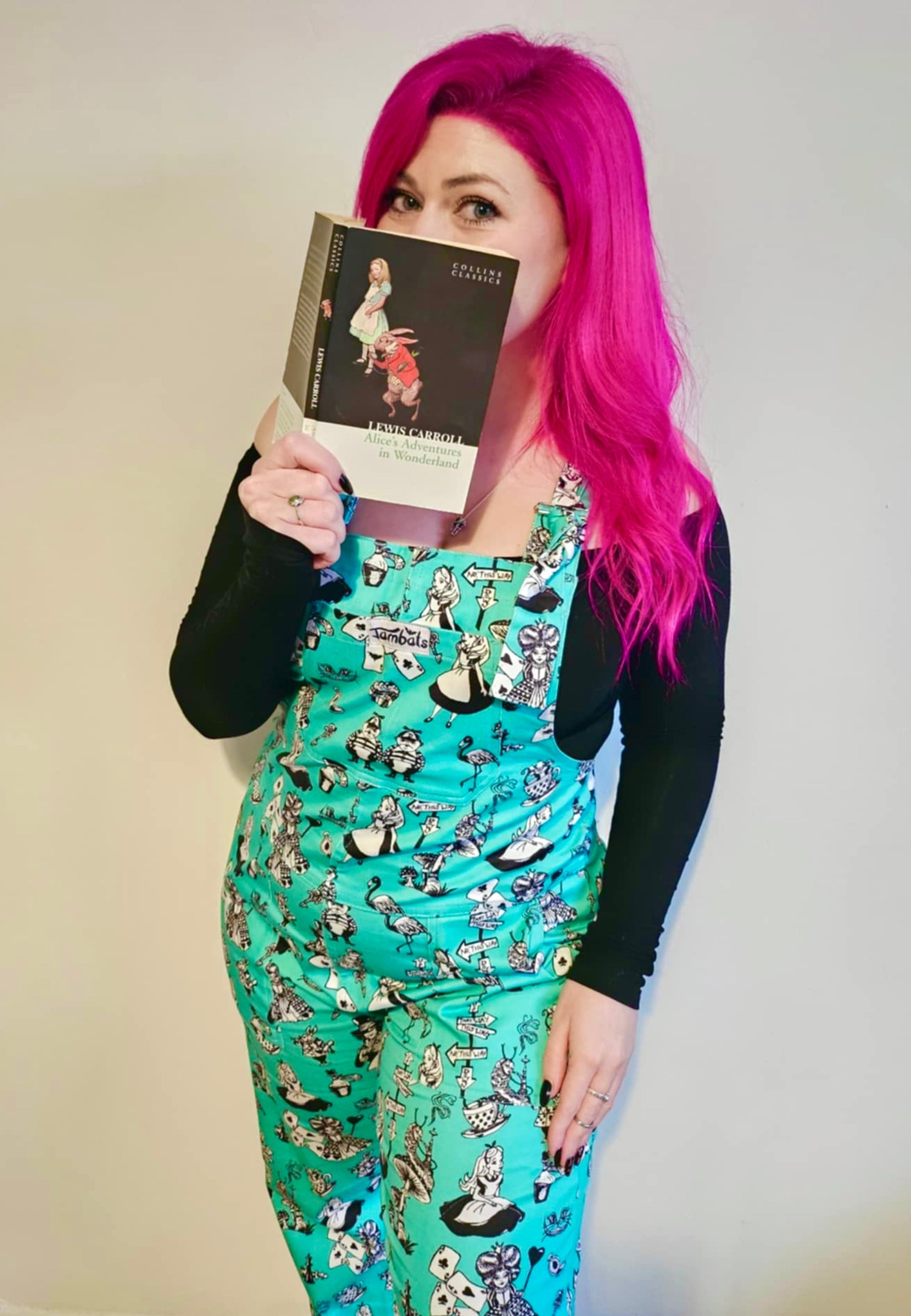 A woman with beautiful deep pink hair I’d wearing Alice in Wonderland print dungarees with black and white characters, standing outdoors.  Woman is cheekily peeping over the tip of an Alice in Wonderland book 