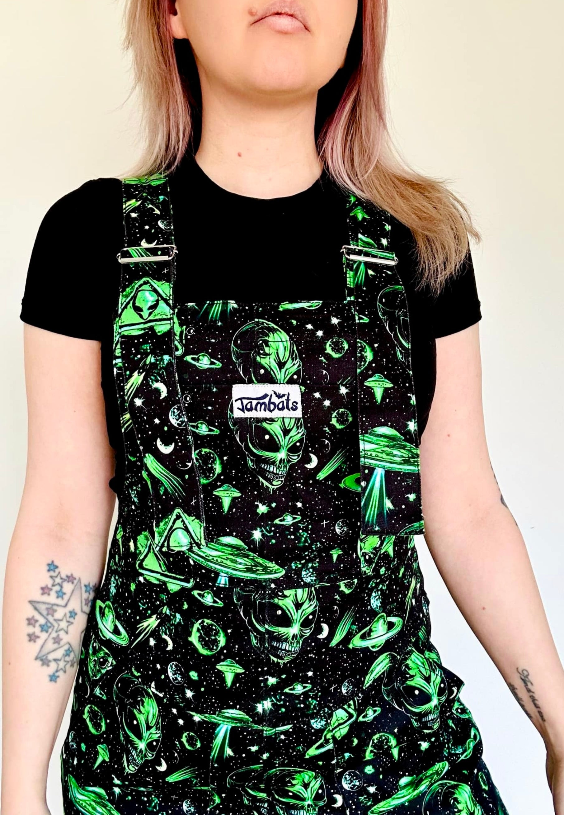 A woman wearing black and green dungarees with an extraterrestrial aliens print standing against a plain background.