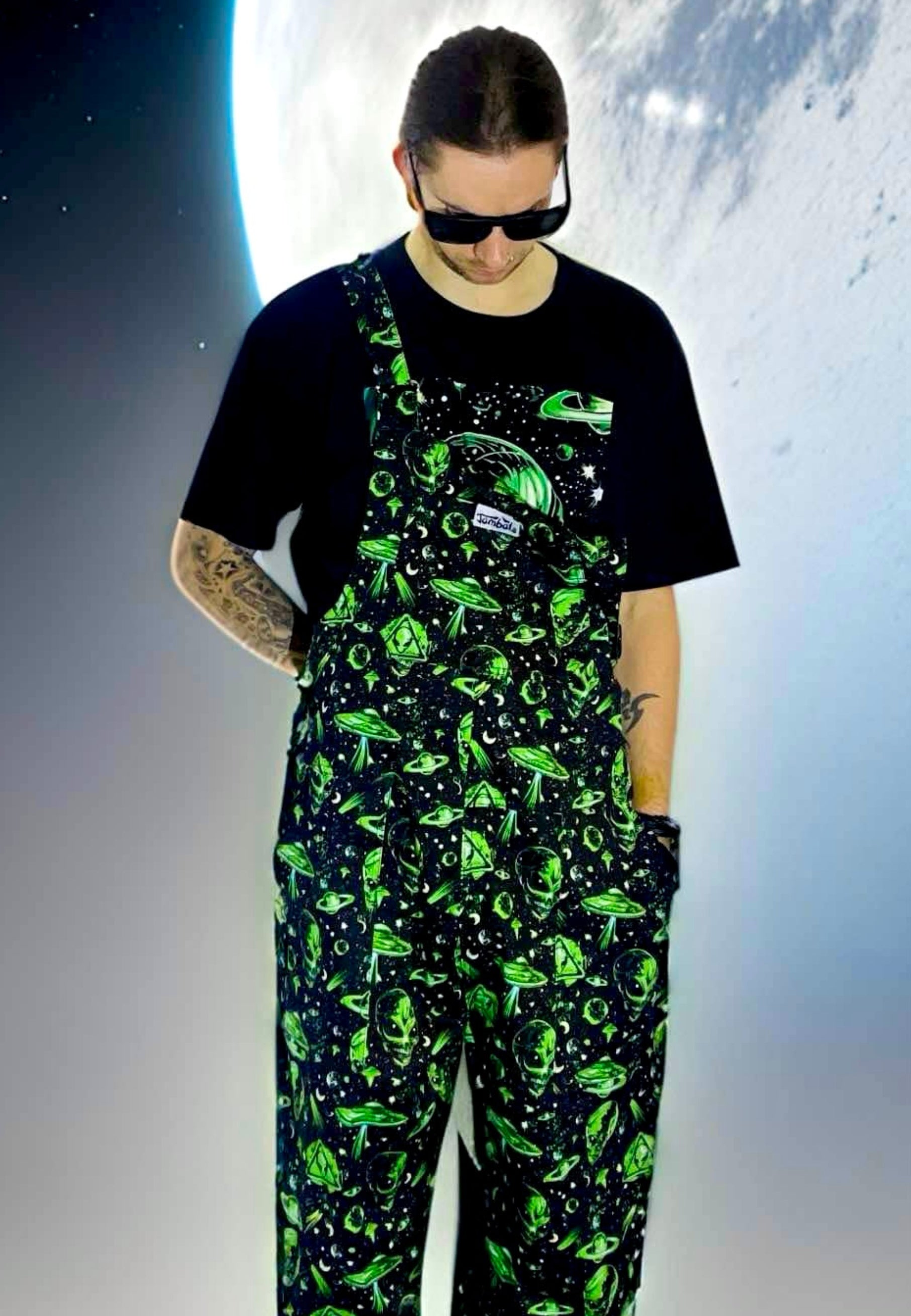 Man wearing black and green dungarees with an extraterrestrial aliens print standing against a space background. Alternative styling 