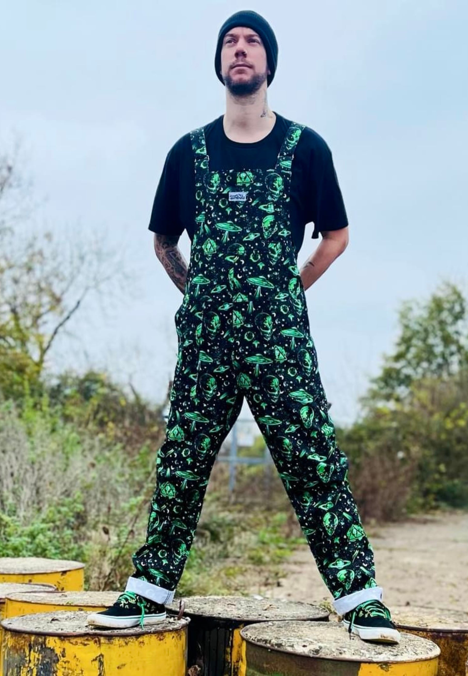 A man wearing black and green dungarees with an extraterrestrial aliens print standing on industrial vats barrels in an area 51 style setting. Alternative styling 