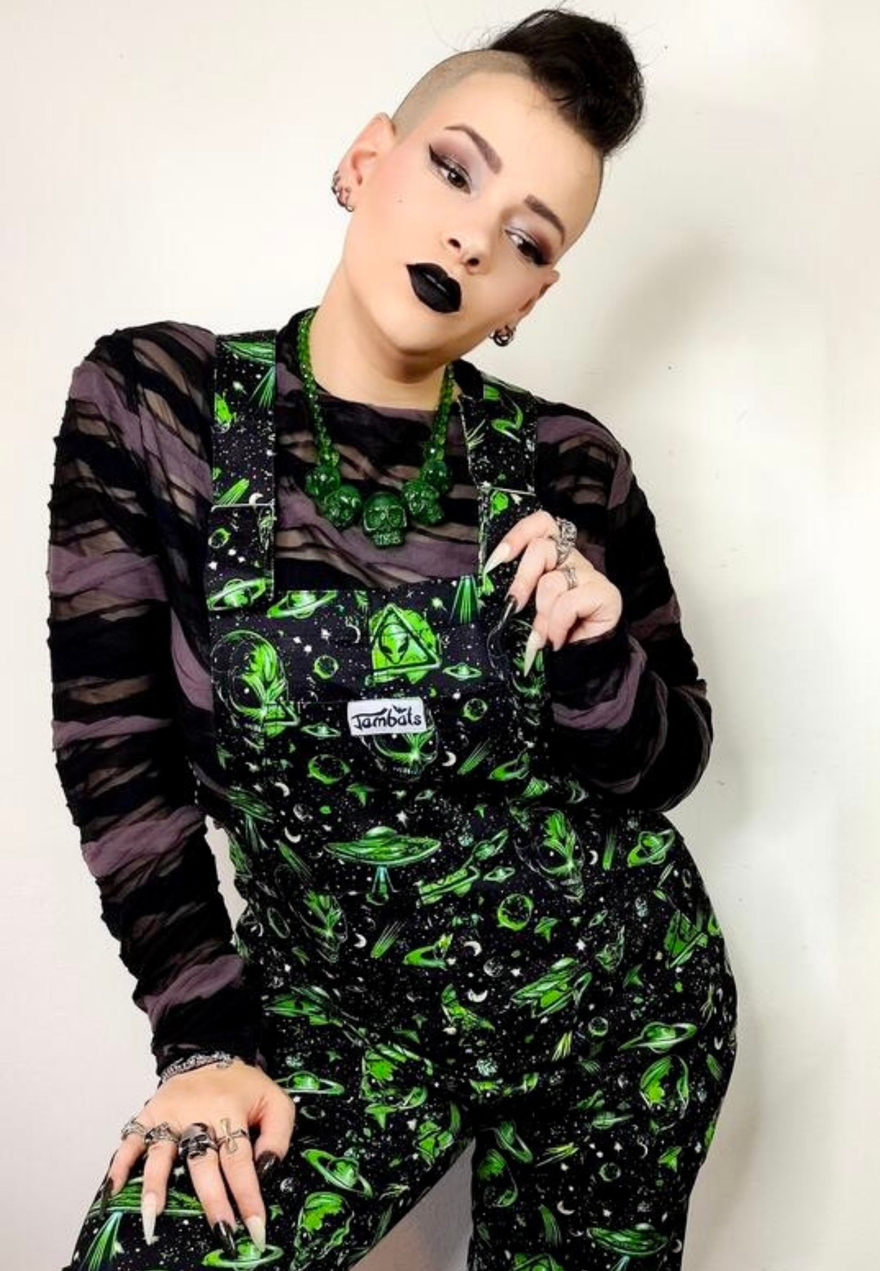 A woman wearing black and green dungarees with an extraterrestrial aliens print standing against a plain background. Alternative punk goth styling 
