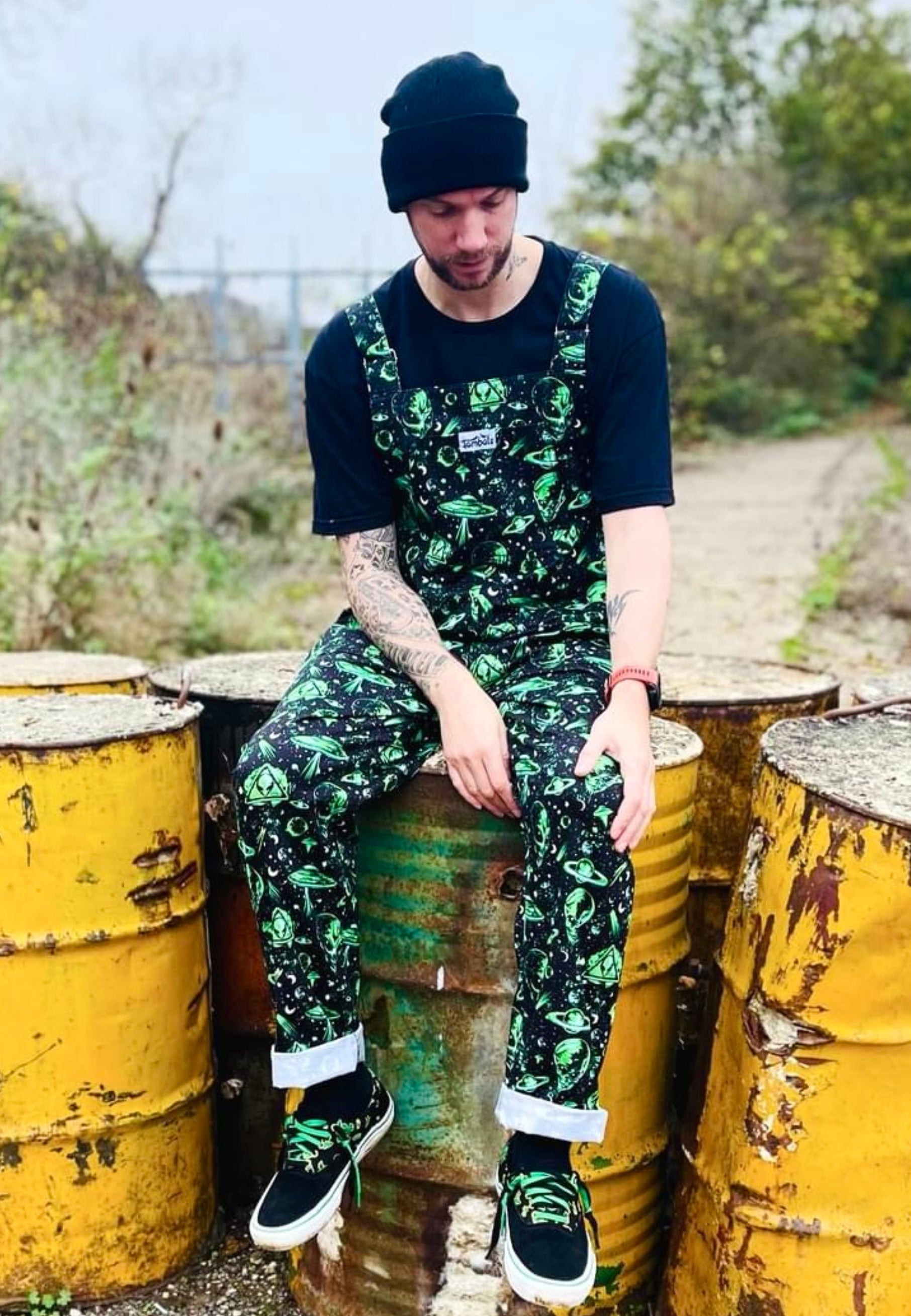 A man wearing black and green dungarees with an extraterrestrial aliens print sat on industrial vats barrels in an area 51 style setting. Alternative styling 