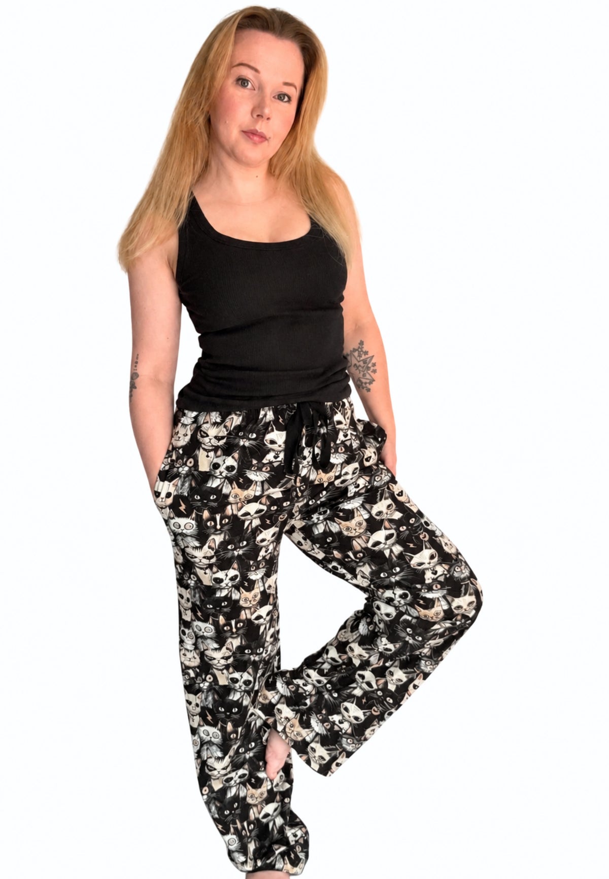 A woman modelling a pair of black and wide leg lounge pants with a beige and white alley cats print, paired with a black tank top, against a plain white background.