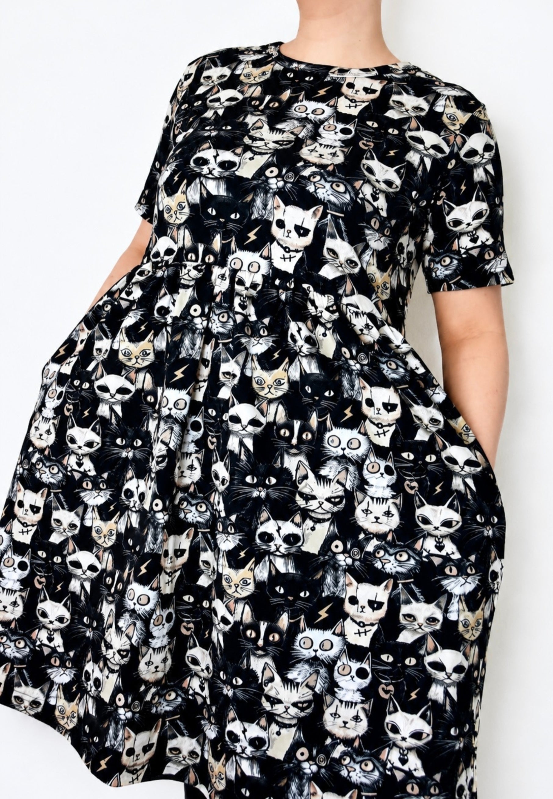 A short sleeve smock dress with a black background and an all over print of stylized punky cat faces in white, gray, and beige, shown on a model against a plain white background.