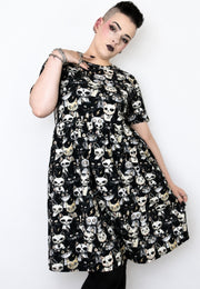 A woman modelling a black smock dress covered in a repeating white and beige punky alley cat print against a plain light background.