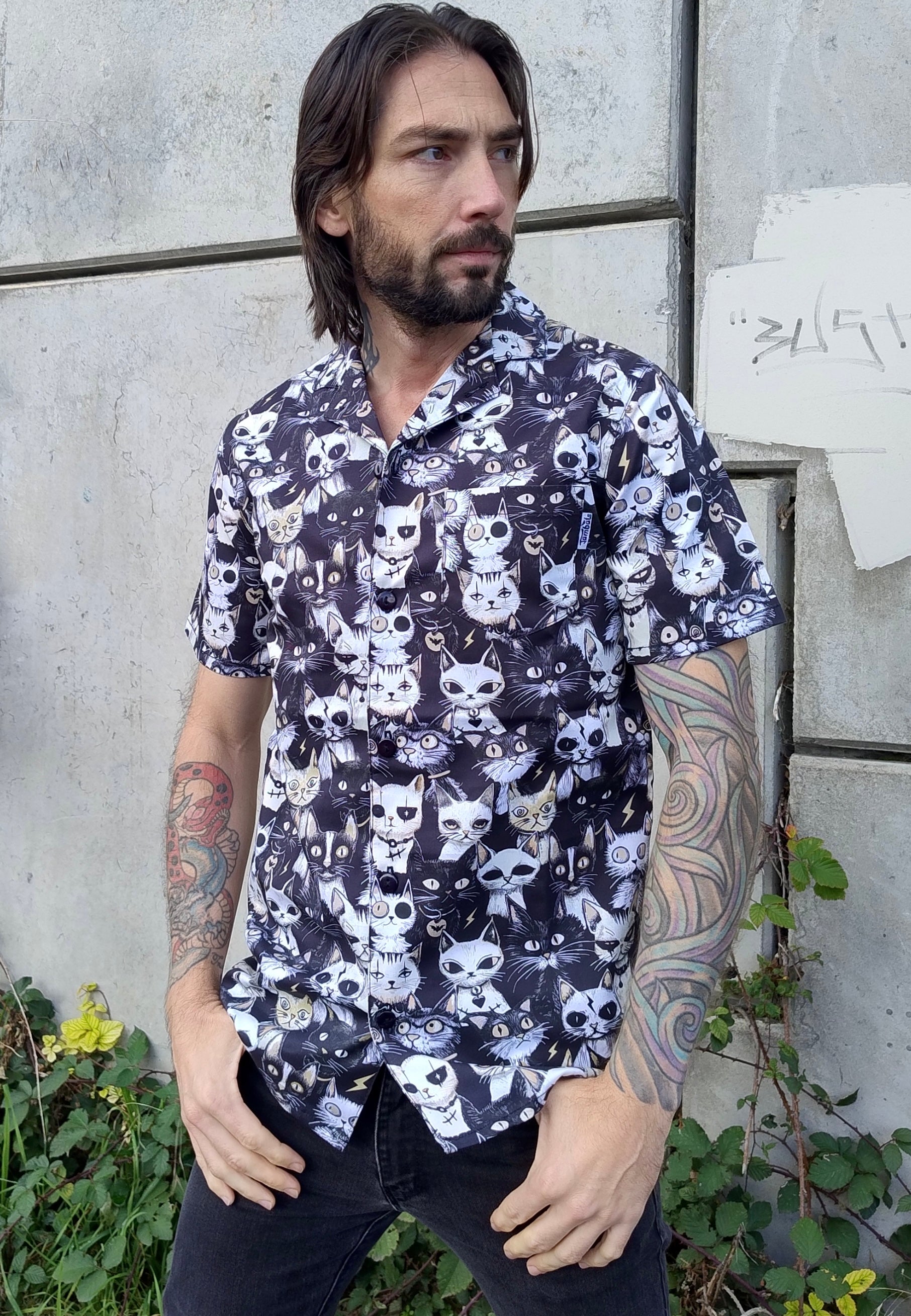 Man wearing a shirt with a print of quirky gothic cats. Outdoor photo against a concrete wall.