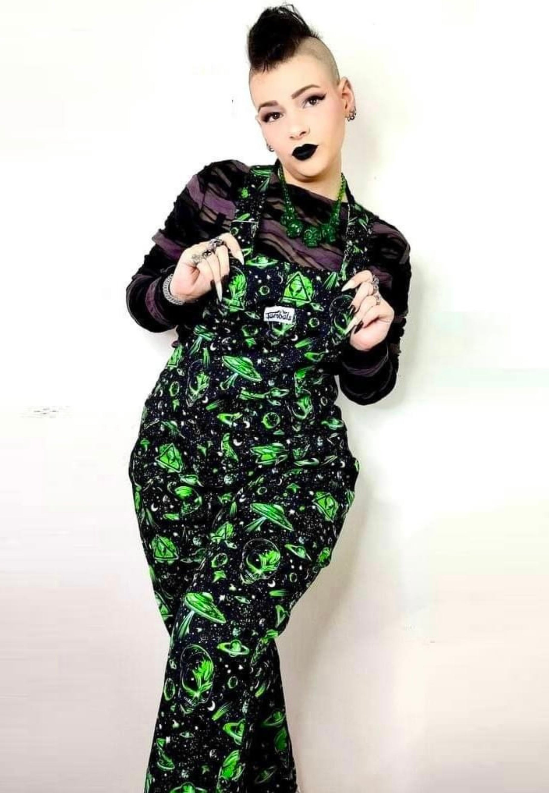 A woman wearing black and green dungarees with an extraterrestrial aliens print standing against a plain background. Alternative punk goth styling 