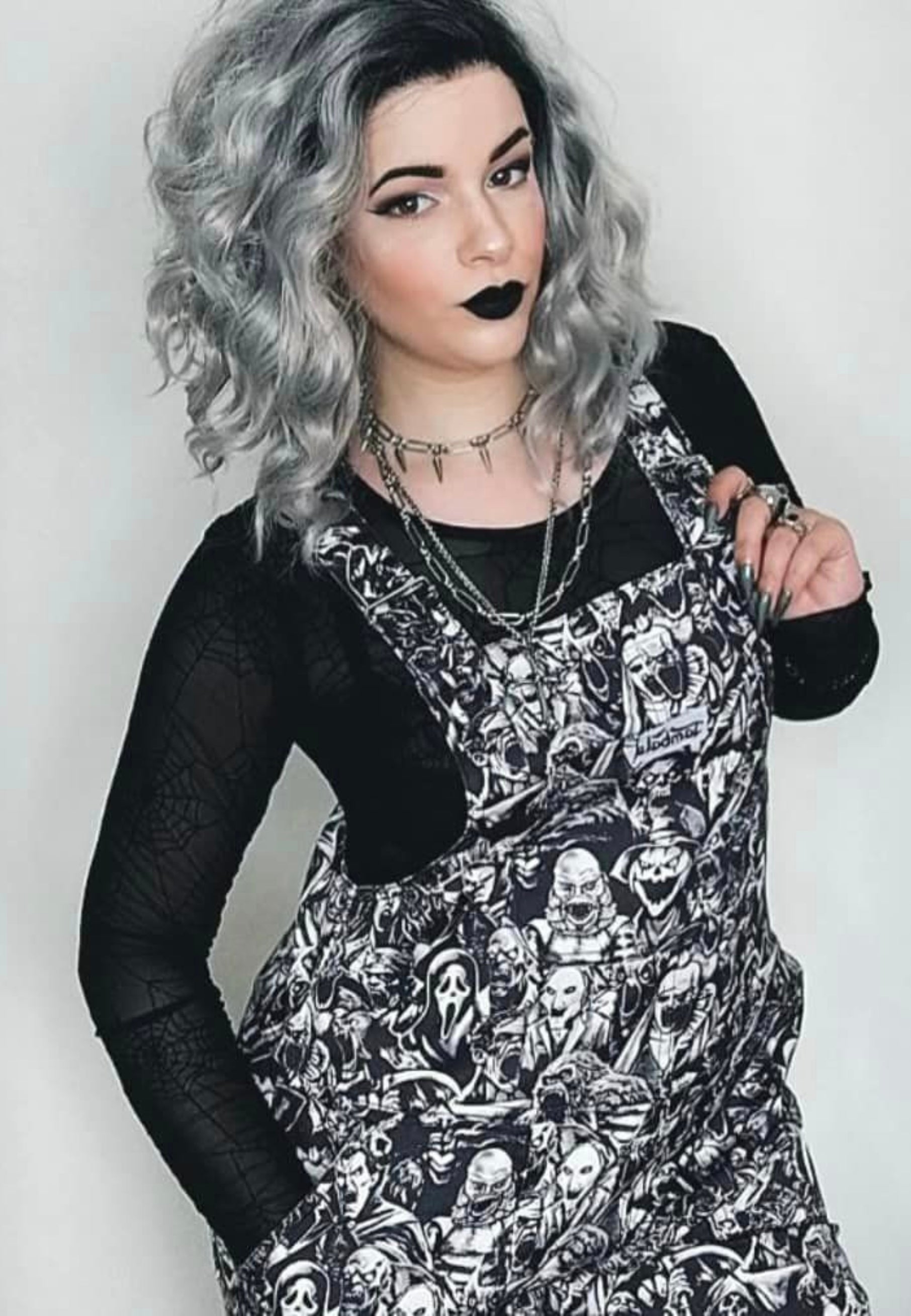 An alternative styled woman  wearing black and white grayscale print dungarees over a black top. Dungaree print is old school and modern monsters, horror characters and cryptids in an old horror comic style illustration. They are all screaming.