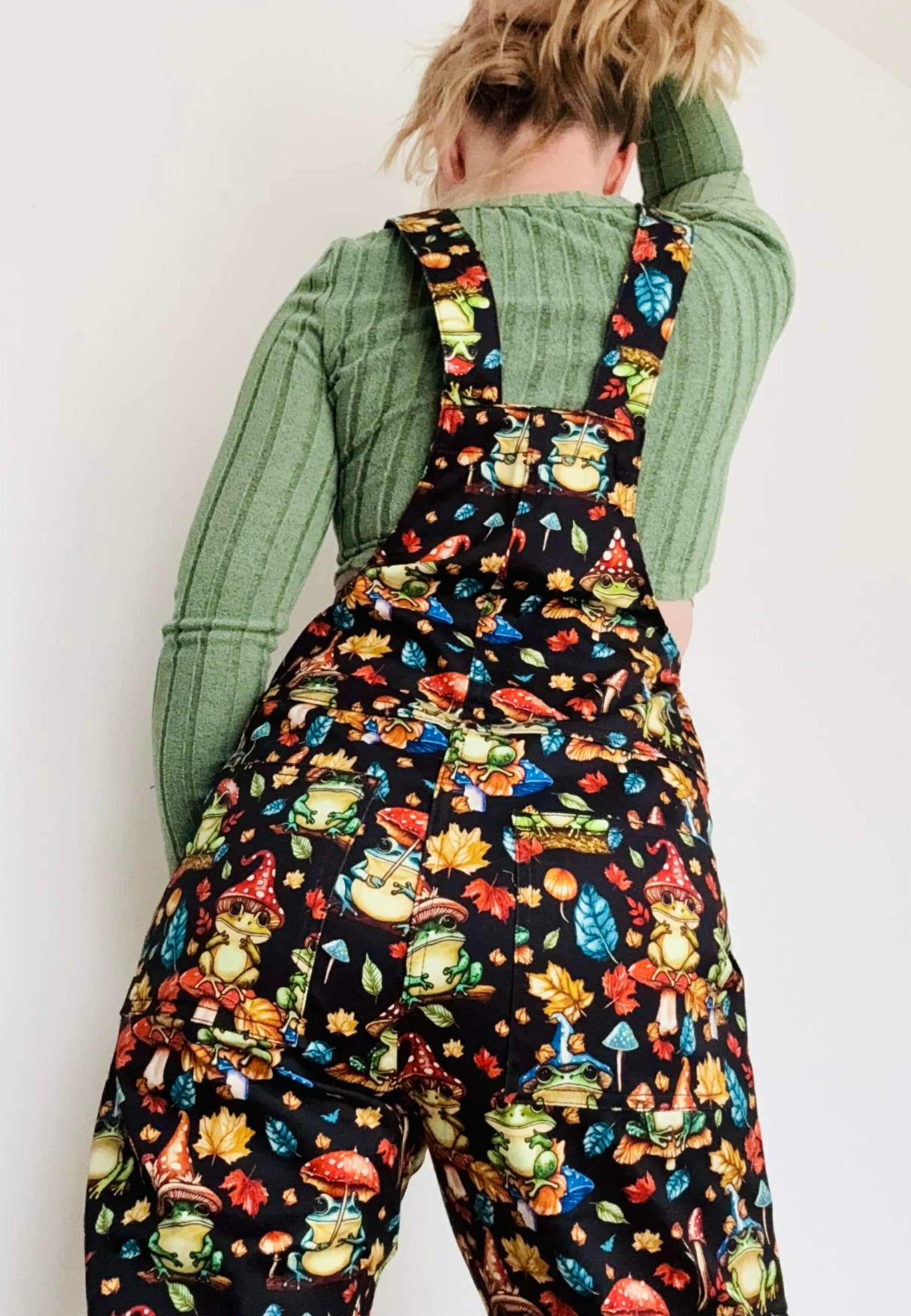 Back view of woman wearing dungarees with a whimsical print of forest frogs with toadstool hats and toadstool umbrellas. Studio photo. 