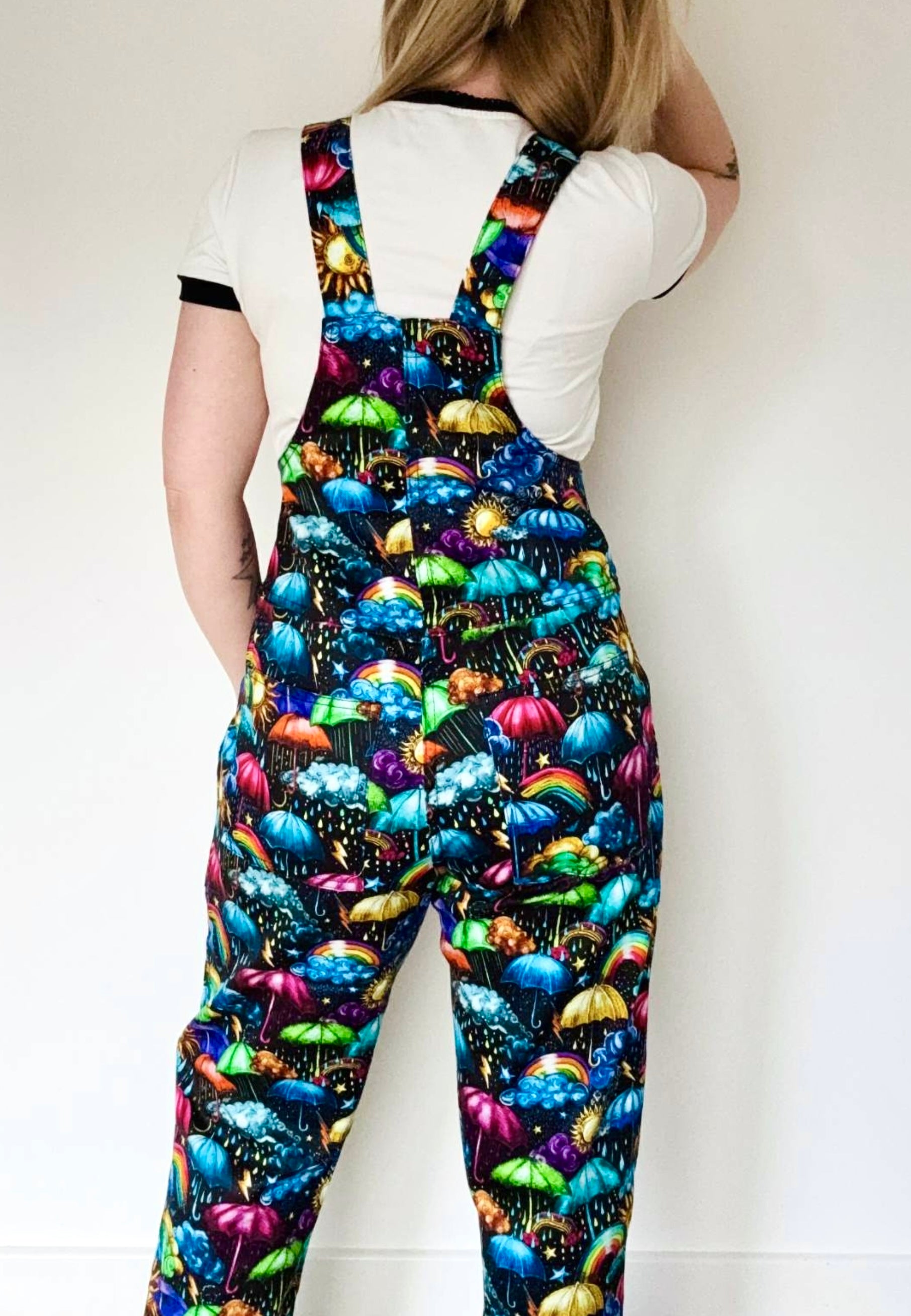 Back view of a woman wearing dungarees with a vibrant weather print. Whimsical fabric print of umbrellas, clouds, rain, sunshine, rainbows, storm clouds. Studio photo. 