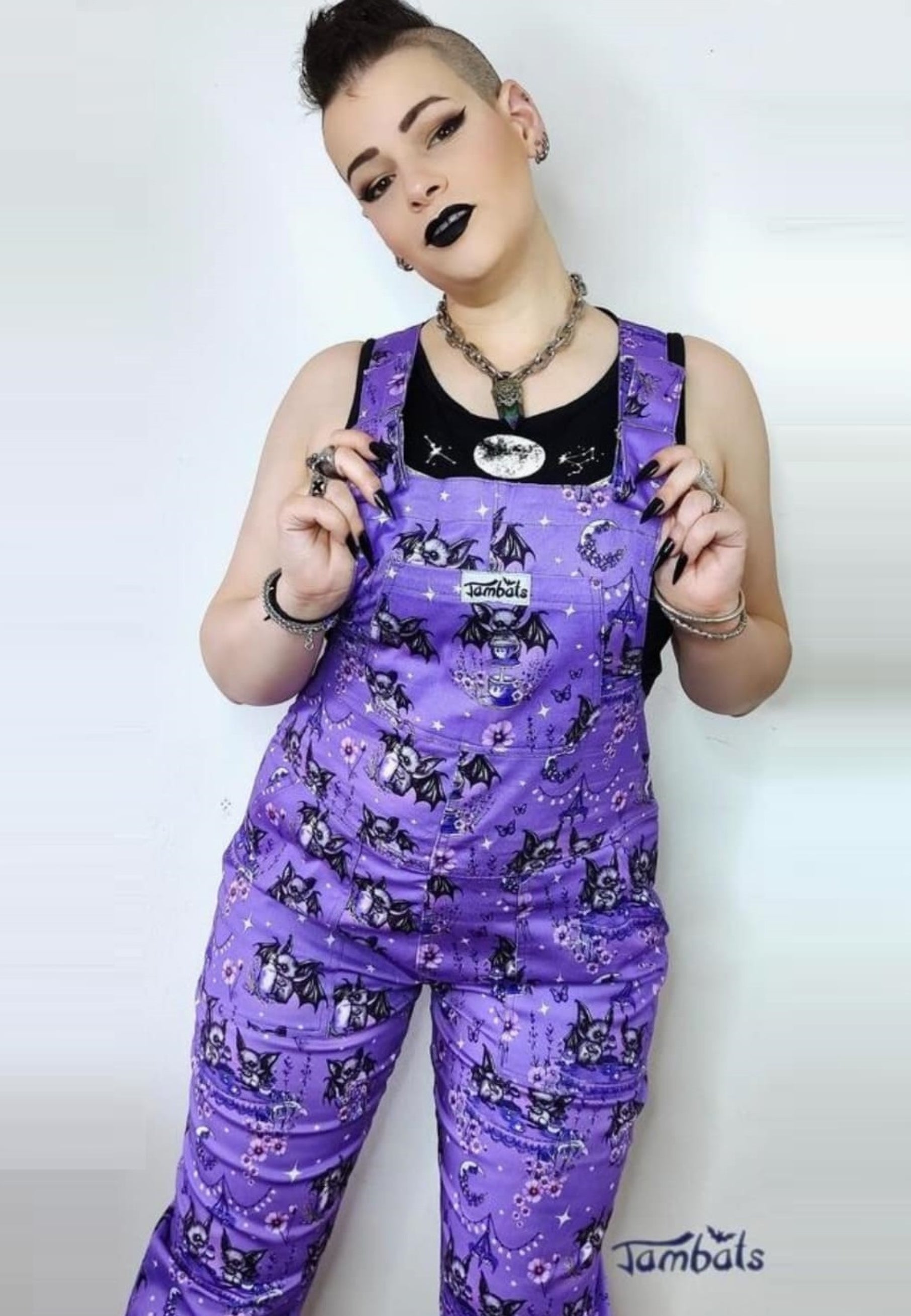 A woman wearing a deep lavender/soft purple dungarees with a cute gothic bats tea party print, paired with a black top. Cute alternative style. 