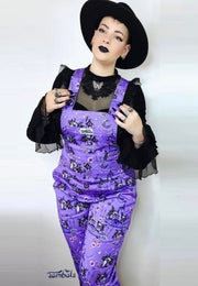 A woman wearing a deep lavender/soft purple dungarees with a cute gothic bats tea party print, paired with a black top and a black hat. Alternative style. 