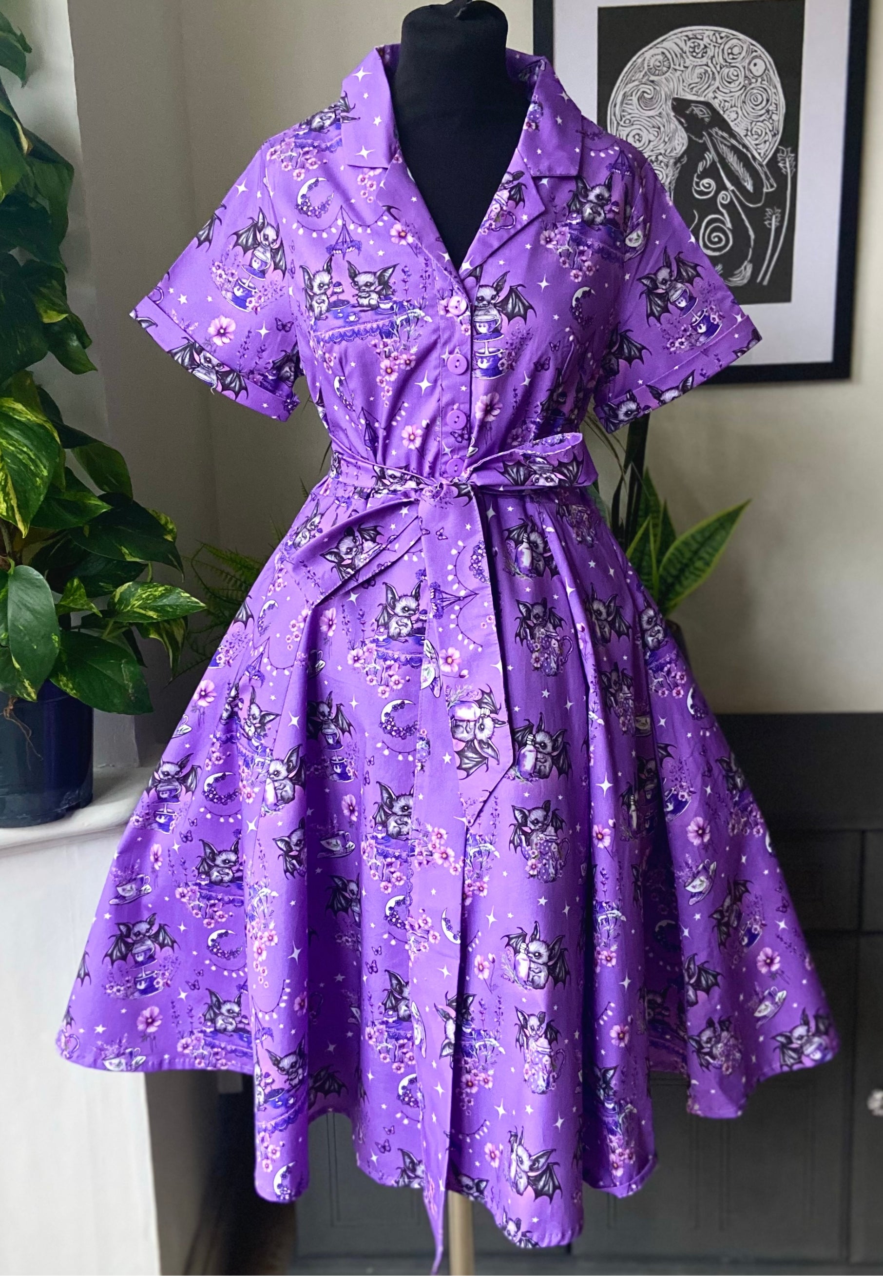 A vintage style purple dress with a beautiful print of little bats having a tea party , featuring a button up blouse, full skirt and three-quarter sleeves. Hourglass shaped dress. Dress is displayed on a mannequin. 