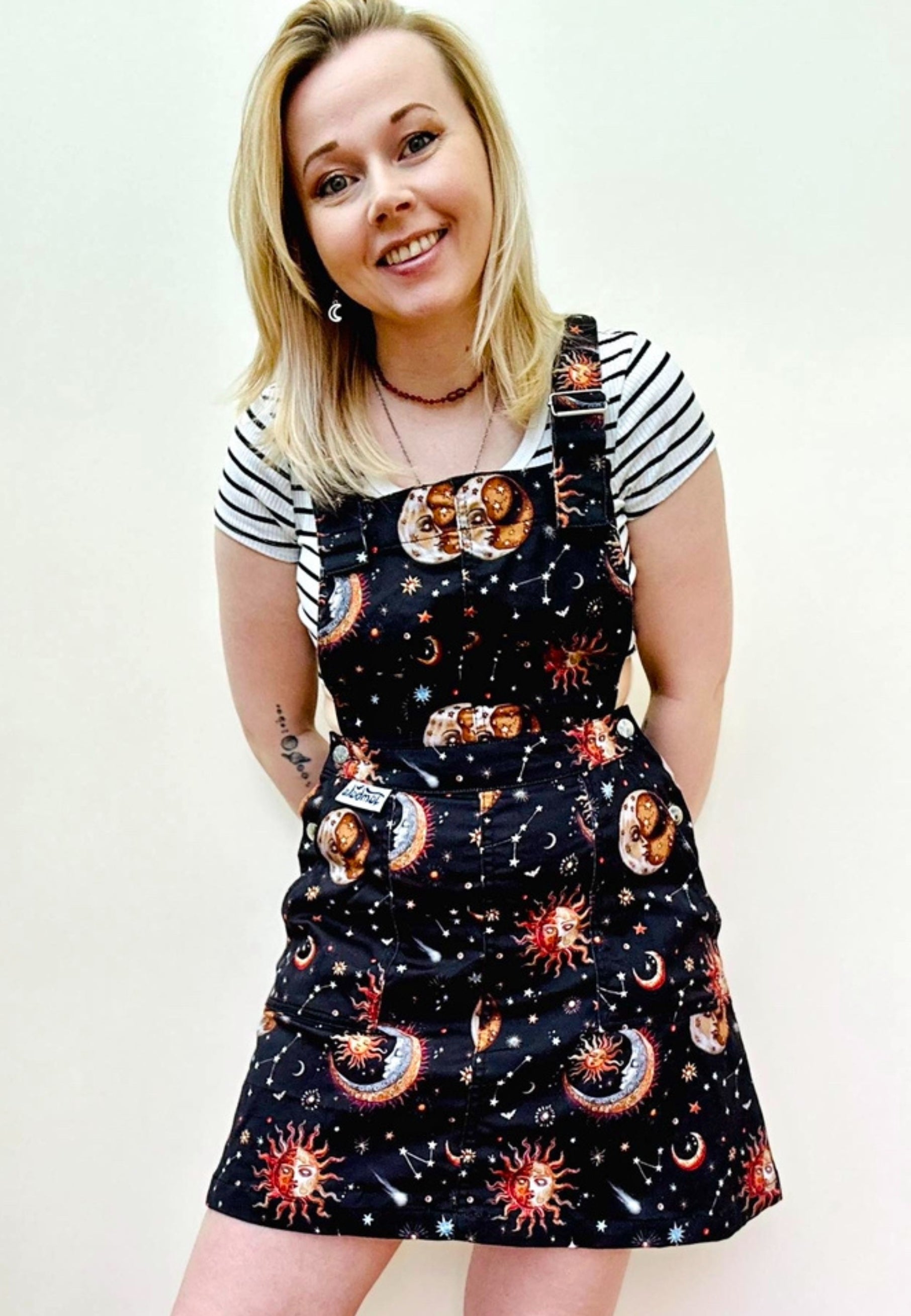 A woman wearing a black pinafore dress with a beautiful celestial print, featuring stars, planets, and moons, paired with a striped top.  