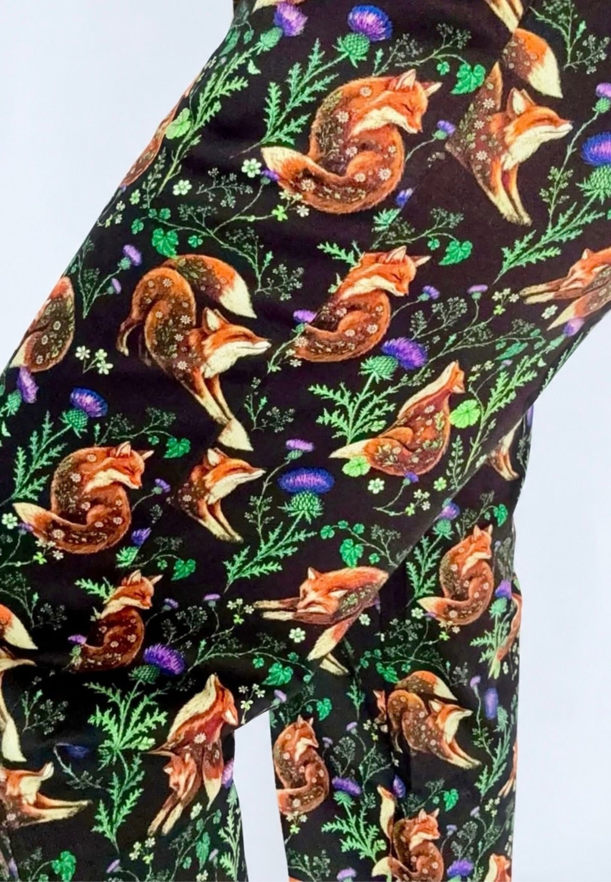 Close up of a pair of trousers featuring an all over print of brown orange foxes, green foliage, and purple thistles on a black background.