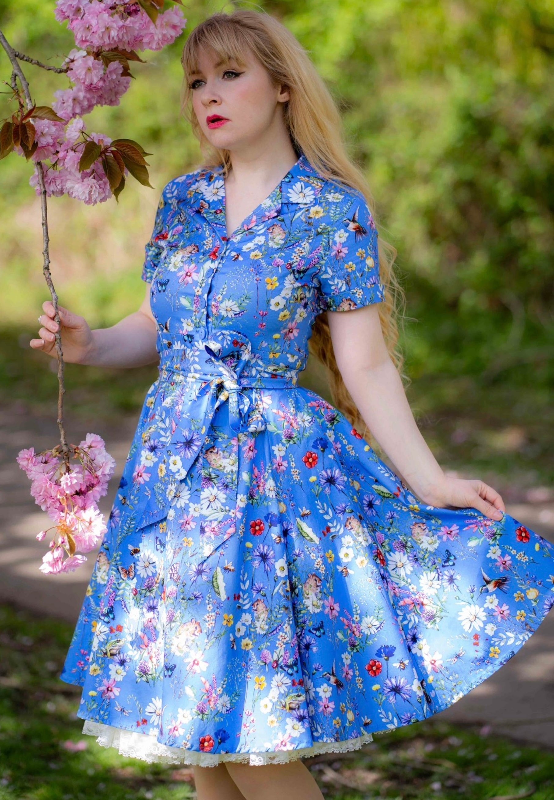 Woman with very long blonde hair wearing a beautiful blue floral dress with a wild meadows print standing outdoors with cherry blossoms.