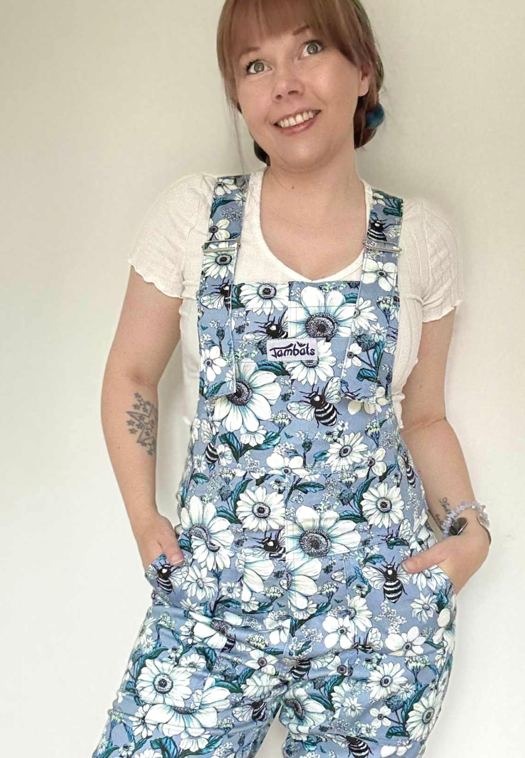 Woman wearing a floral daisy and bees patterned dungaree shorts against a plain background. JamBats dungaree shorts. JamBats shorts. overalls. 