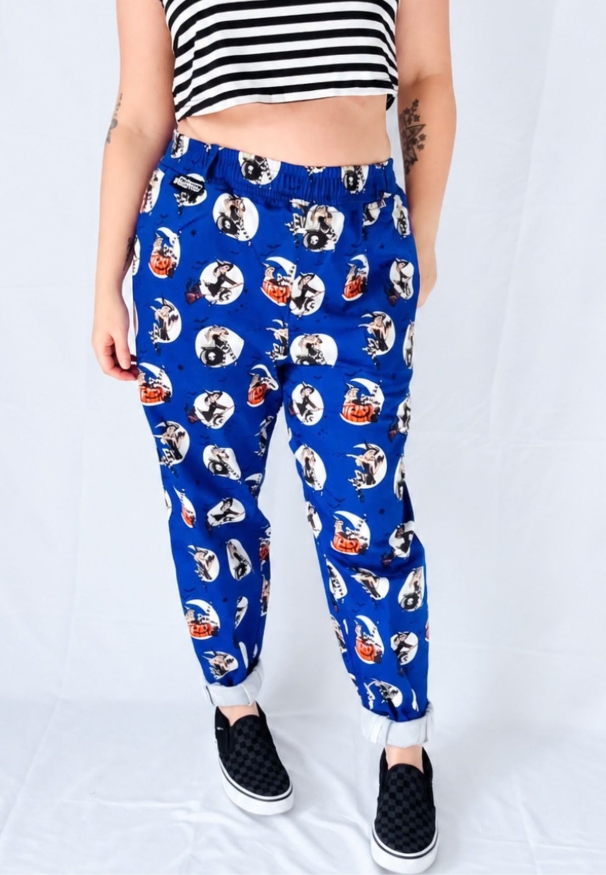 A woman modelling a pair of blue trousers with an all over pattern of spooky retro pin up witches, paired with a black and white striped crop top, against a white studio background
