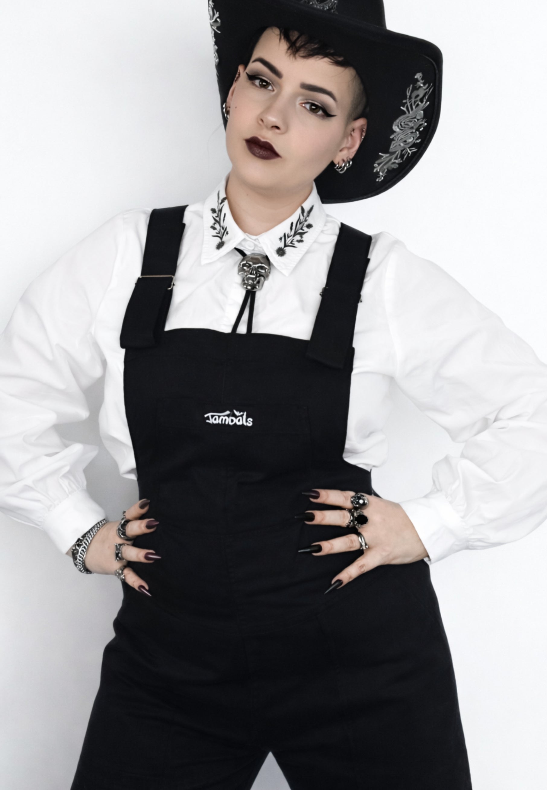 Woman wearing a black cowboy hat, white shirt, and black dungarees with a white background. Alternative style
