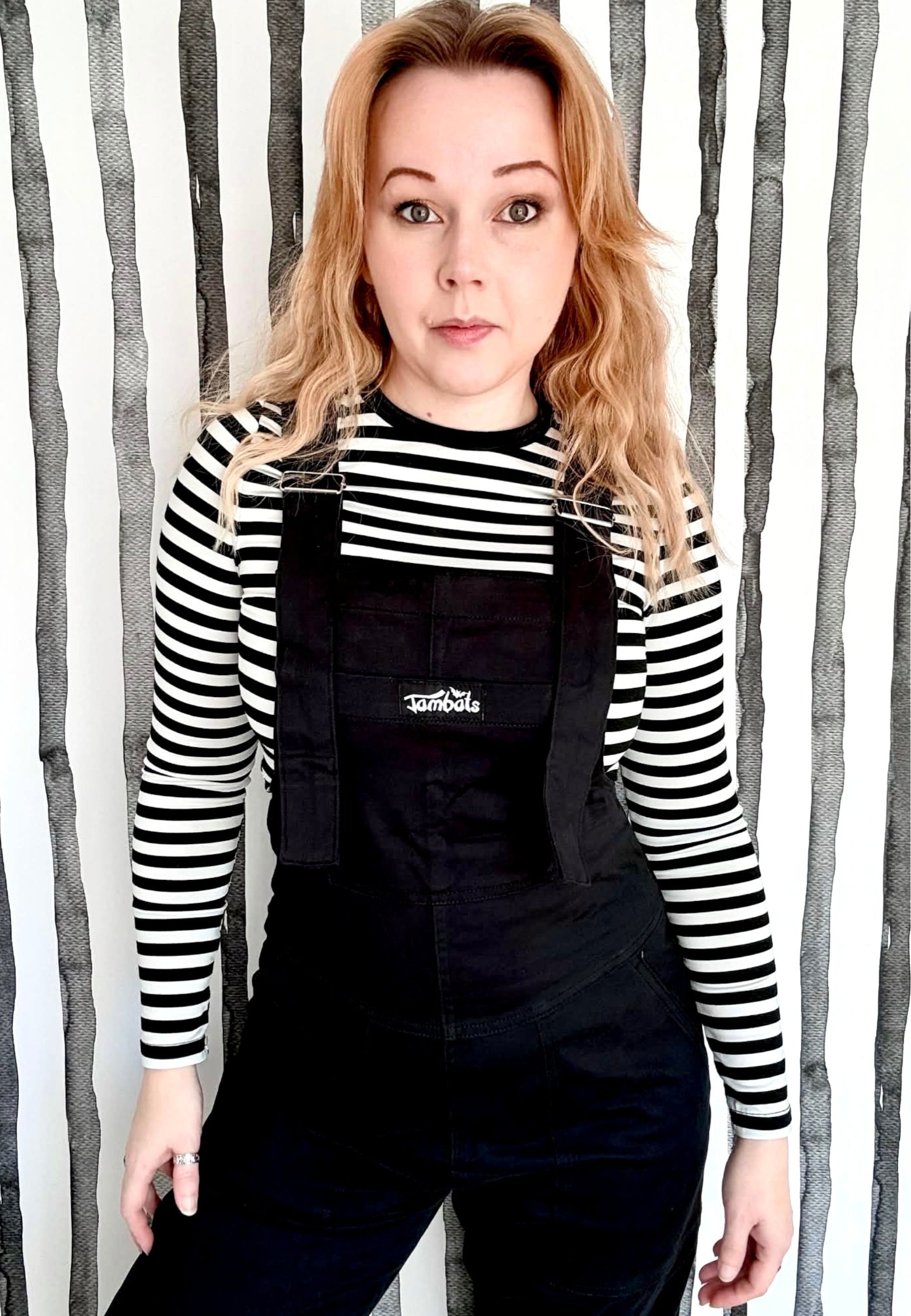 Woman wearing black dungarees overalls and a black and white striped top against a quirky striped wall.