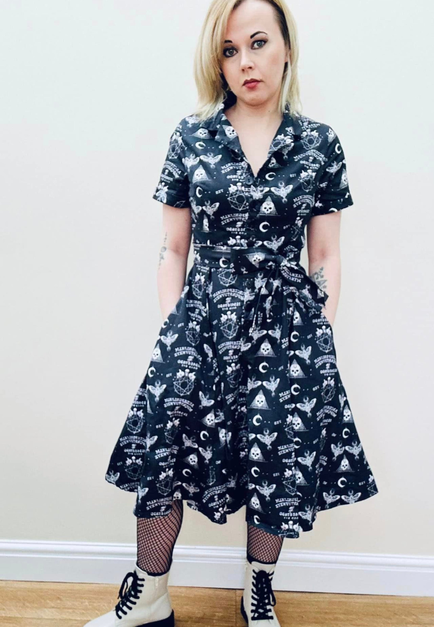 A woman wearing a black gothic Ouija print dress with a full skirt and button bodice. Styled with boots and black fishnet tights. 