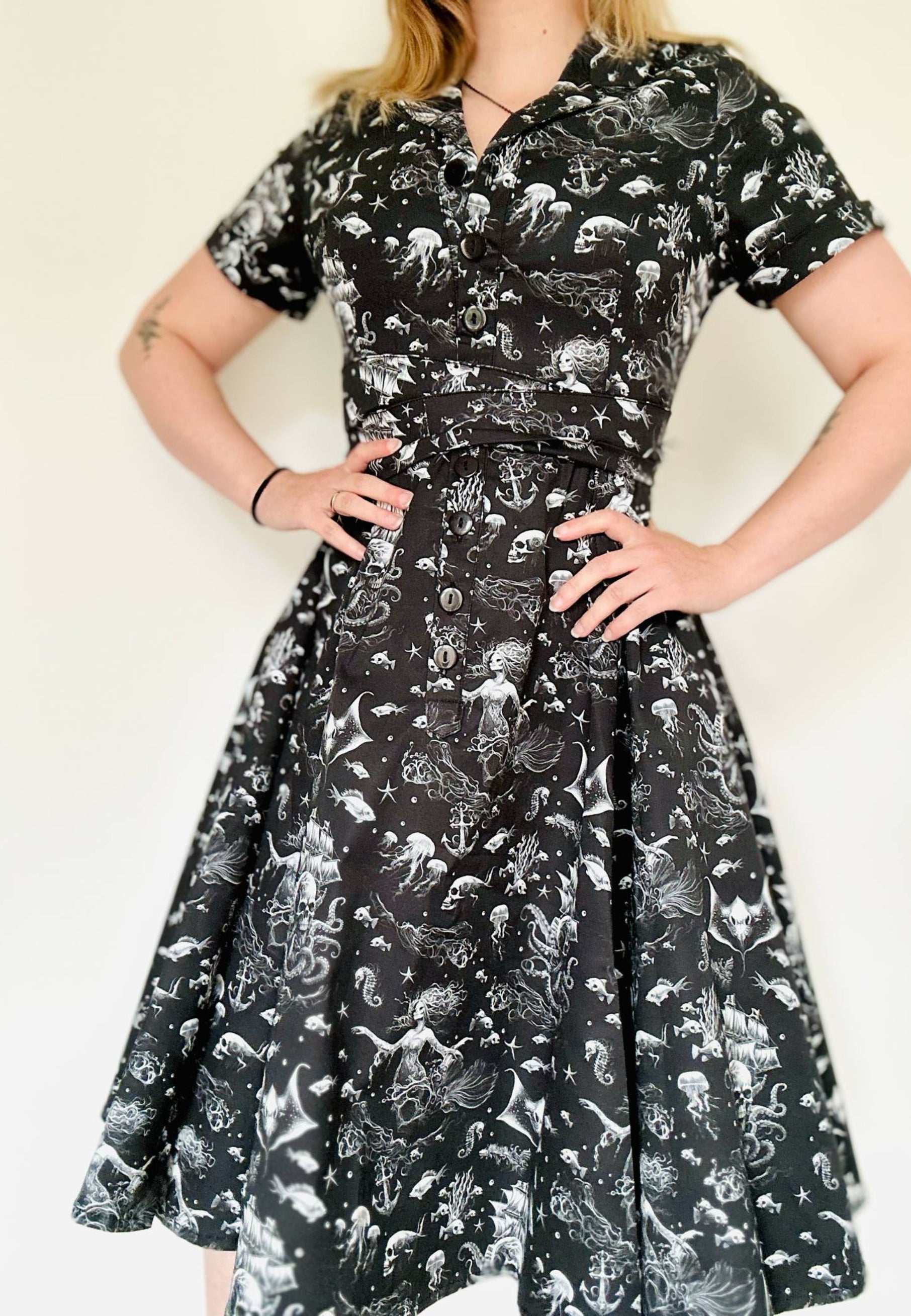 Woman wearing a black shirt dress with a beautiful gothic print of under the sea. Ghostly sirens and sunken skull pirate ships. Studio photo against a plain background. Goth style dress.