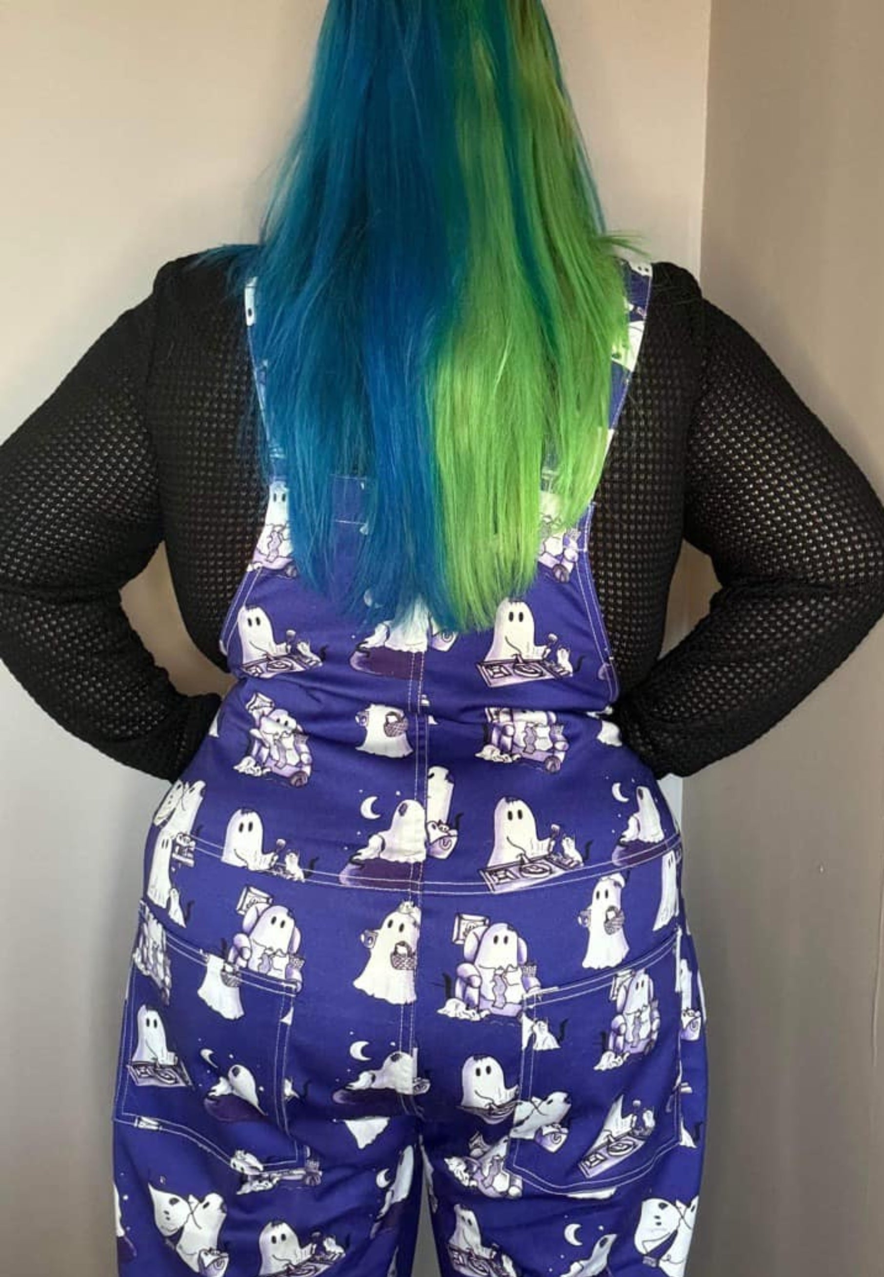Back view of a witchy woman in a witches hat wearing cute blue dungarees with a print of ghosts and ghost cats. 