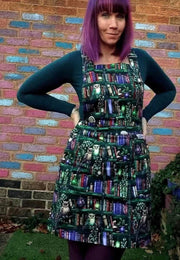 A woman wearing a green pinafore dress with a unique bookcase and owls print, standing in front of a colorful brick wall.