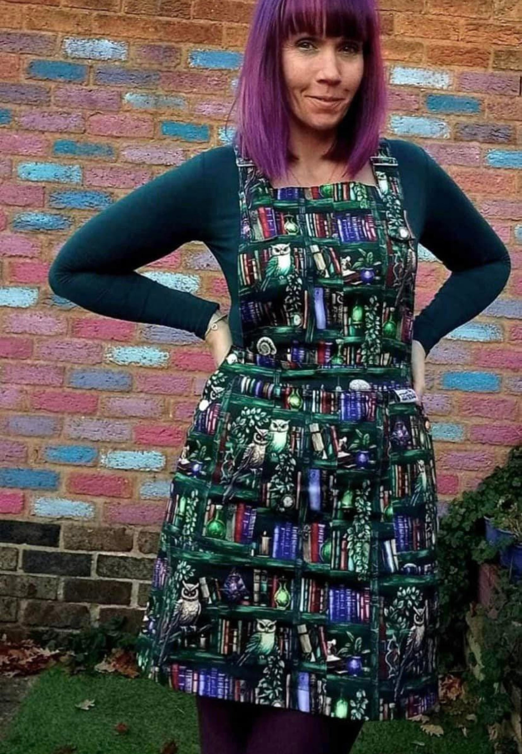 A woman wearing a green pinafore dress with a unique bookcase and owls print, standing in front of a colorful brick wall.