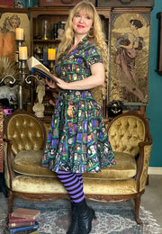 A woman stands in a vintage room wearing a beautiful shirt dress with a colorful book case print pattern, purple and black striped tights, and black boots. She is posed on a gold velvet chaise lounge in front of a bookshelf and framed artwork.