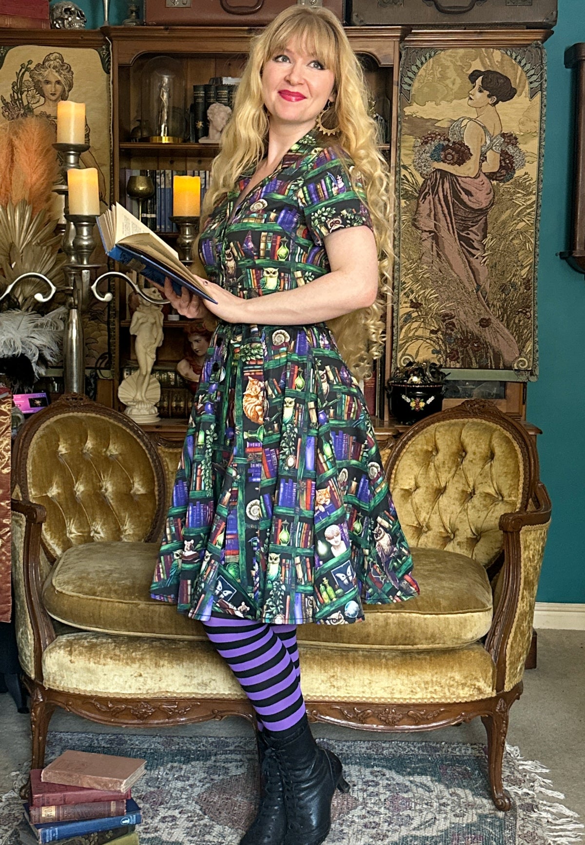 A woman stands in a vintage room wearing a beautiful shirt dress with a colorful book case print pattern, purple and black striped tights, and black boots. She is posed on a gold velvet chaise lounge in front of a bookshelf and framed artwork.