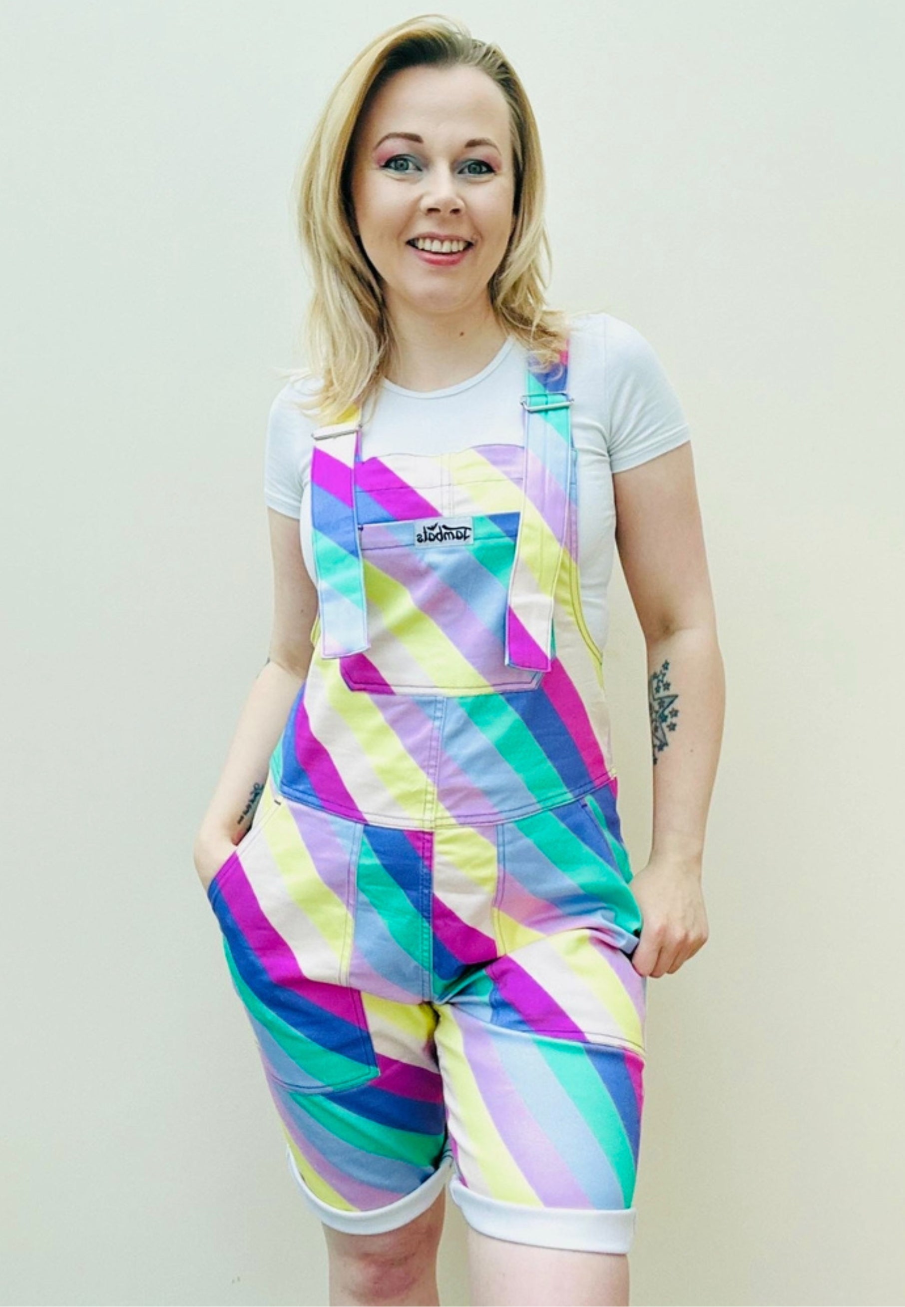 A person wearing bright multicolored striped shorts dungarees with a chest pocket, standing against a plain background. Bright pastel striped dungaree shorts. Retro style. 