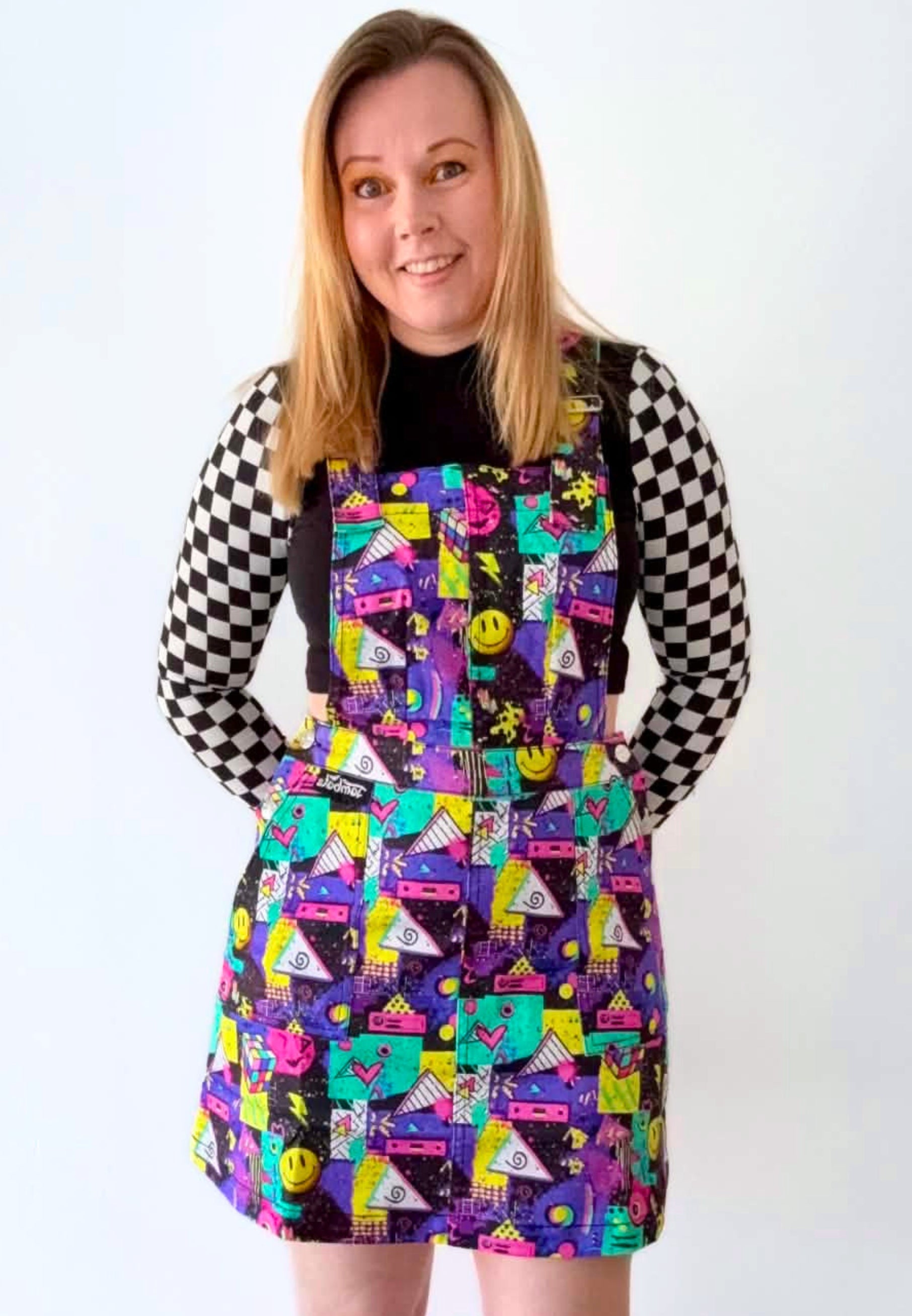A woman modelling a colourful retro themed pinafore dress with a vibrant  80s 90s pattern of smiley faces, cassette tapes, and geometric shapes, worn over a black and white checkered long-sleeve shirt against a plain white background.