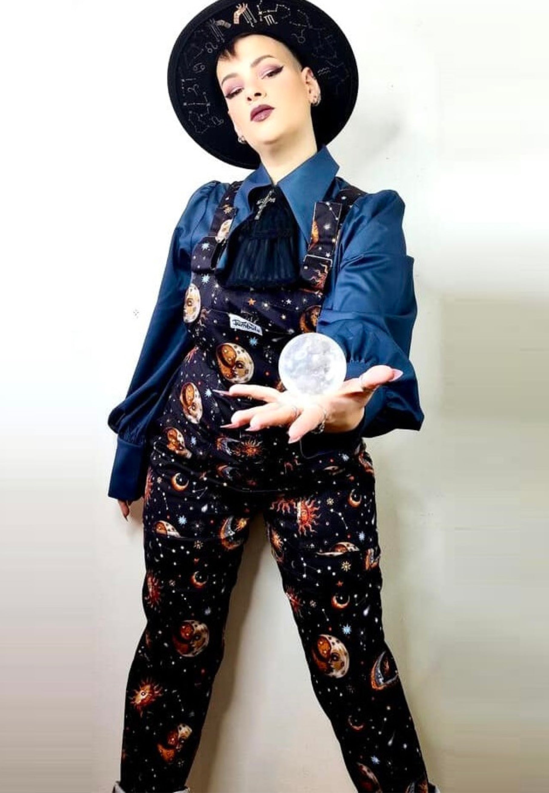 A woman wearing a regular fit, celestial print dungarees with a chest pocket, hip pockets, and a bum pocket. The dungarees are black with a unique fabric print featuring moons and stars. Mystical Witch Style. 
