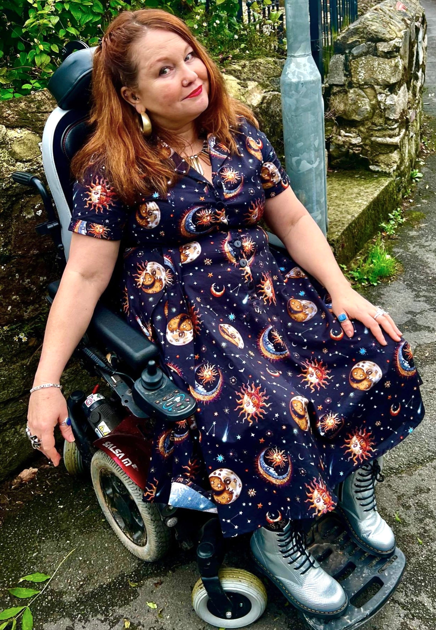 Woman in a wheelchair wearing a beautiful dress with celestial, whimsical sun and moons pattern in an outdoor setting.