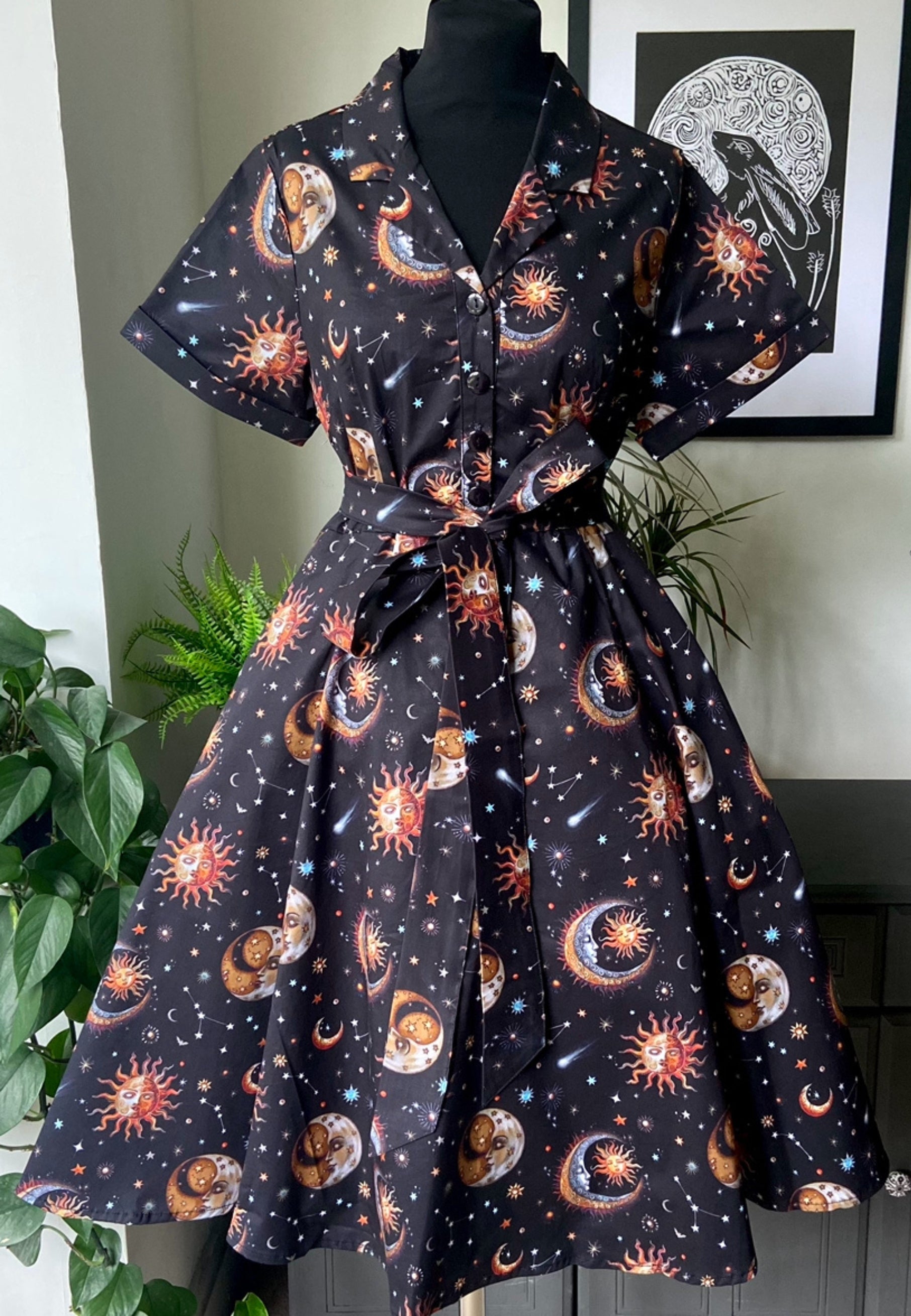Beautiful midi retro dress with a celestial print consisting of moons, stars, and planets on a black background. The dress features a button-up shirt bodice and a tie belt at the waist.
