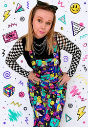 A woman posing in colourful , patterned dungarees overalls over a black checkered long-sleeve shirt, set against a white background decorated with 90s style graphics.