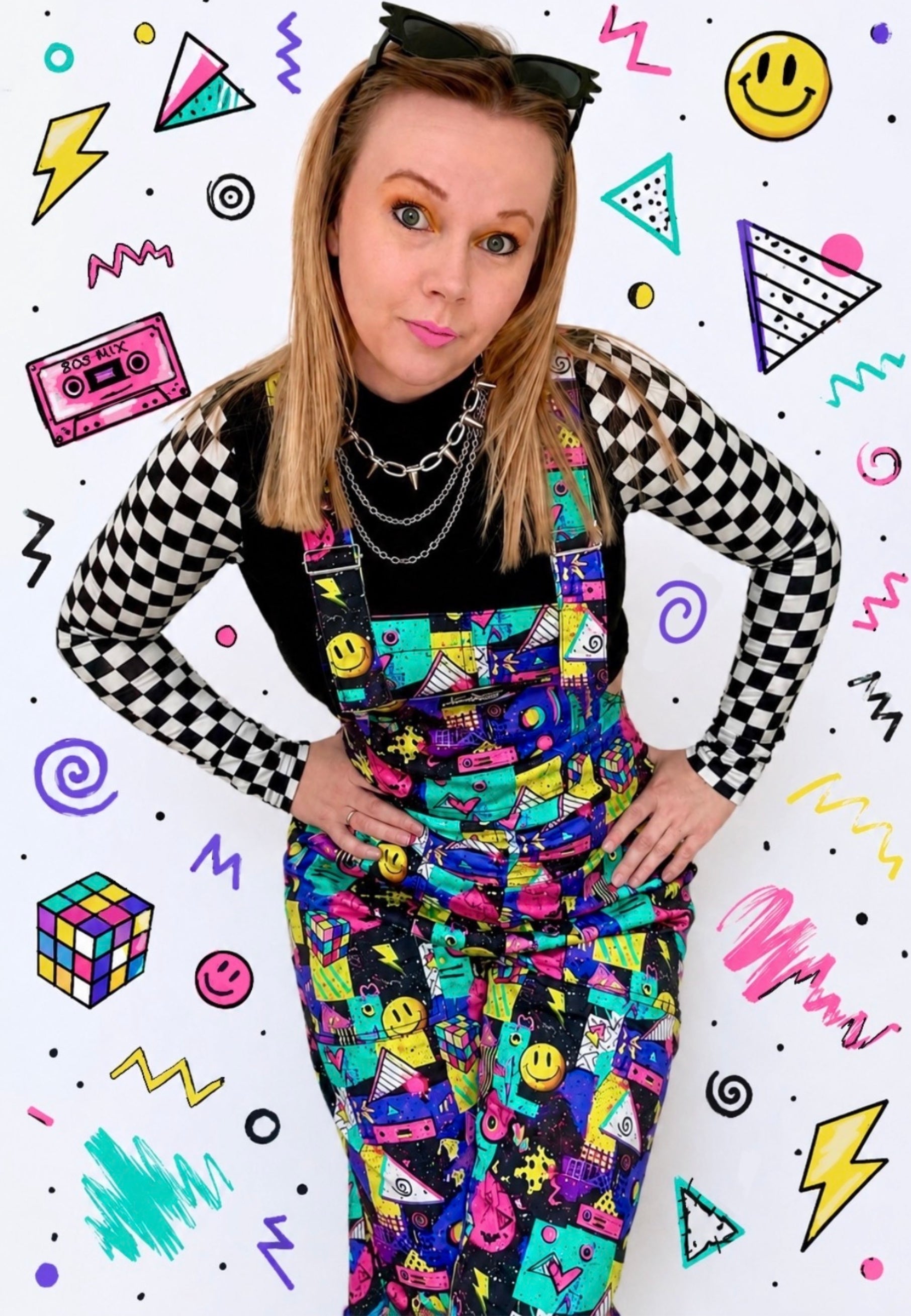 A woman posing in colourful , patterned dungarees overalls over a black checkered long-sleeve shirt, set against a white background decorated with 90s style graphics.