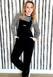 Woman wearing black dungarees overalls and a striped shirt against a quirky striped wall.